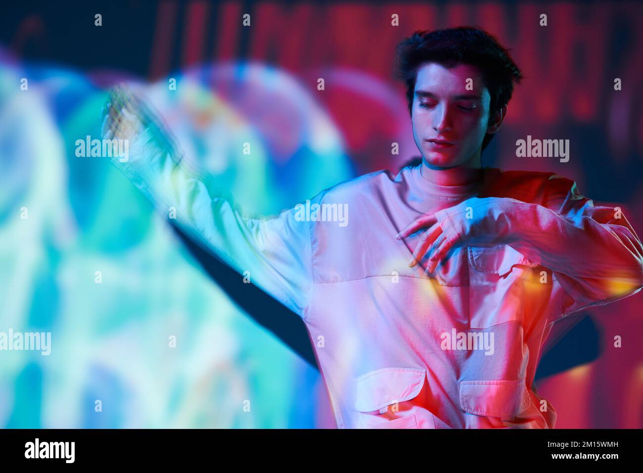 Junger Mann, dunkelhaariges Modell in Sweatshirt mit vielen Taschen, die im Studio stehen, mit ausgestreckten Armen und geschlossenen Augen in leuchtendem Neon, mehrfarbigen li Stockfoto