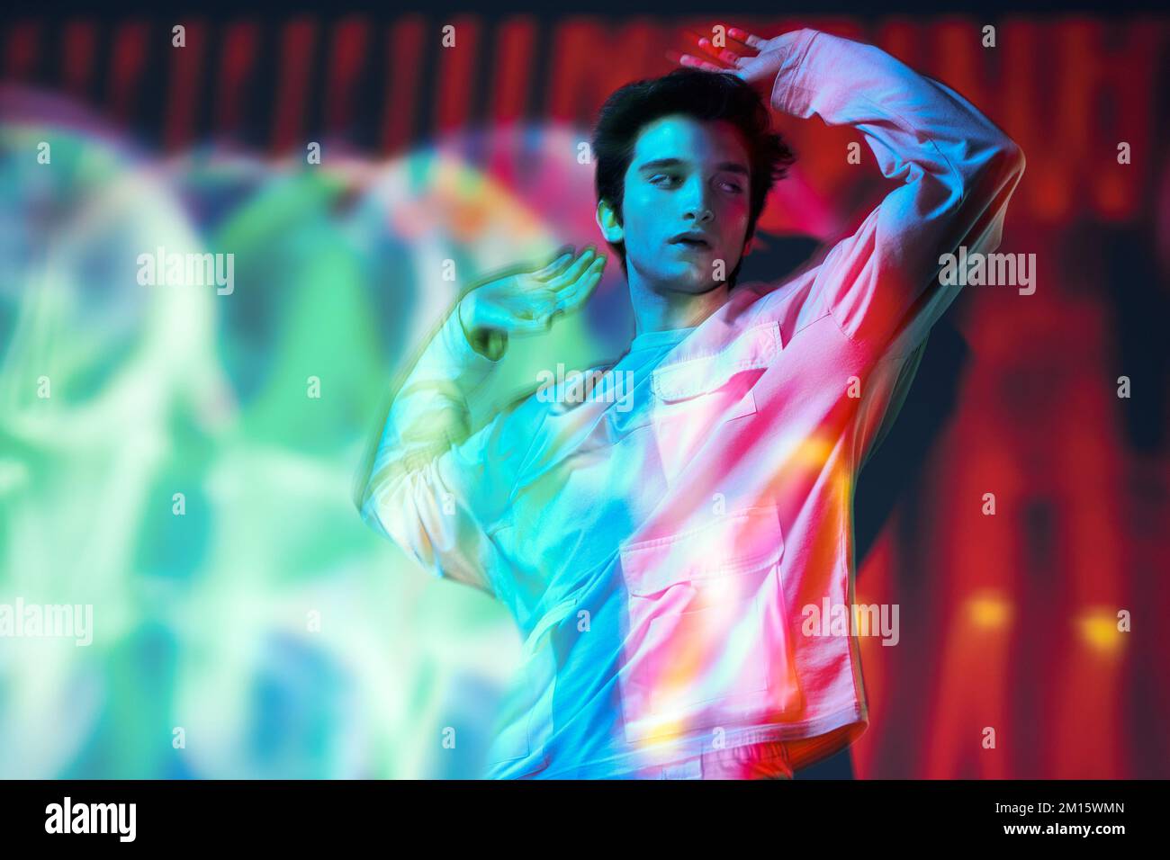 Junger Mann, dunkelhaariges Modell in Sweatshirt mit vielen Taschen, steht im Studio mit ausgestreckten Armen und schaut in leuchtendem Neon-Multikolo auf die Kamera Stockfoto