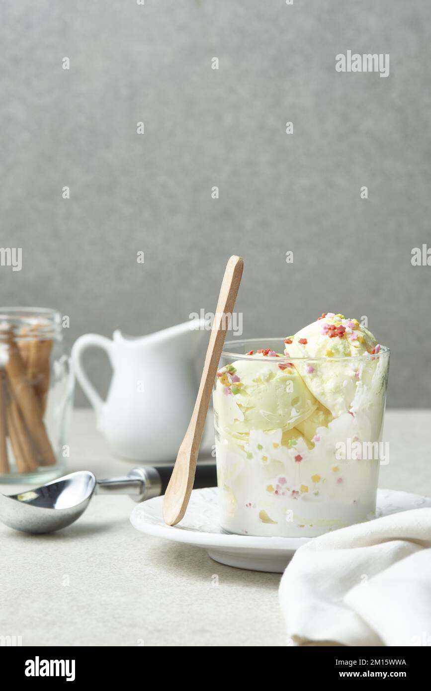 Schaufeln mit köstlichem Vanilleeis mit bunten süßen Streuseln, serviert in einem Glasbecher auf dem Tisch mit einem Holzlöffel aus Zimtstangen und einem Krug Stockfoto