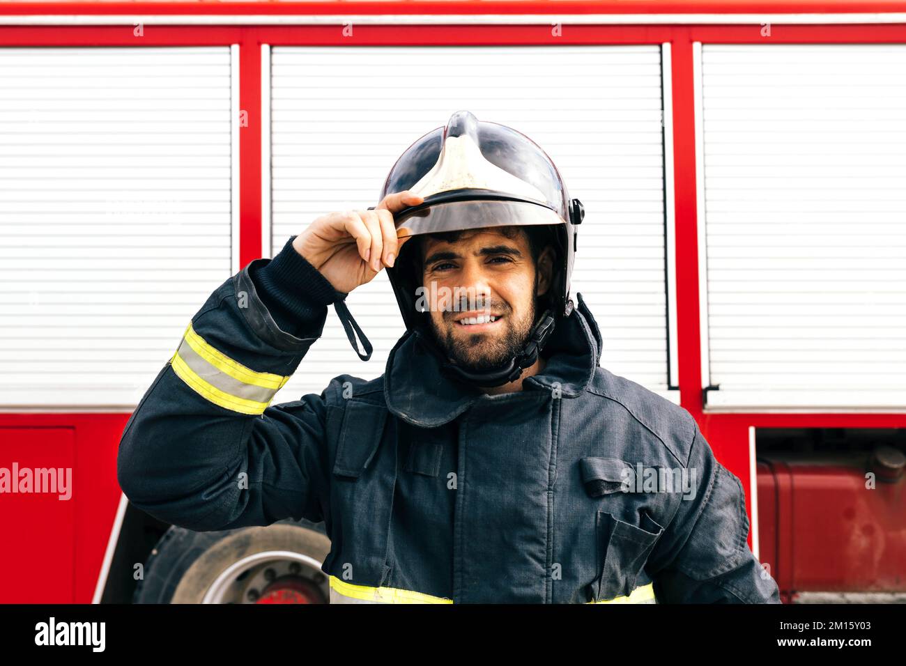 Positiver hispanischer Feuerwehrmann in Schutzuniform, der mit einem Lächeln auf die Kamera schaut und den Helm anpasst, während er neben dem Feuerwehrwagen auf der Station steht Stockfoto