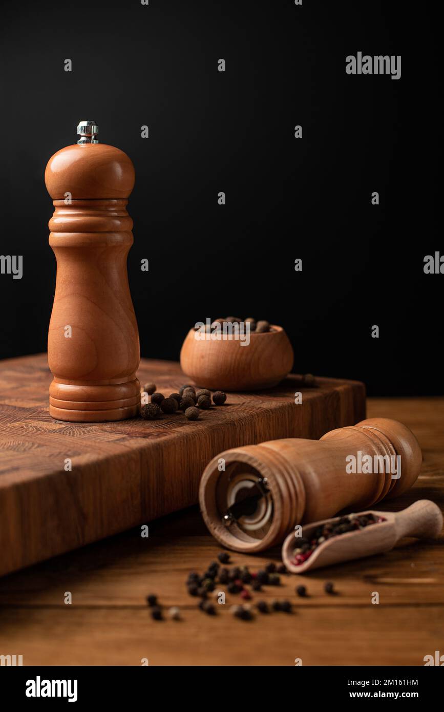 Pfeffermühle und ganzer Pfeffer in Holzschaufel auf schwarzem Tisch. Stockfoto