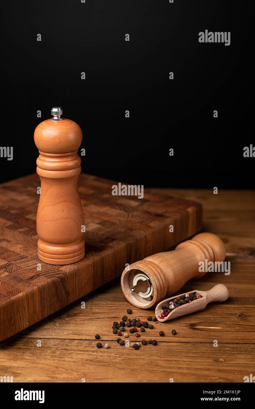 Pfeffermühle und ganzer Pfeffer in Holzschaufel auf schwarzem Tisch. Stockfoto