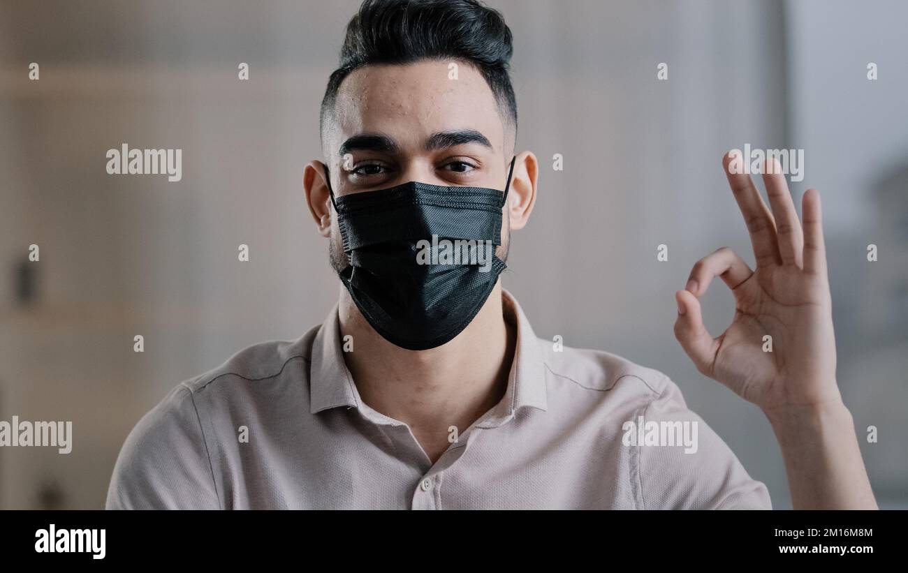 Glücklich hispanischen männlichen Mitarbeiter arabischen Mann Unternehmer tragen medizinische Maske verhindern Coronavirus Epidemie Blick auf Kamera zeigt okay Zeichen alles Fi Stockfoto