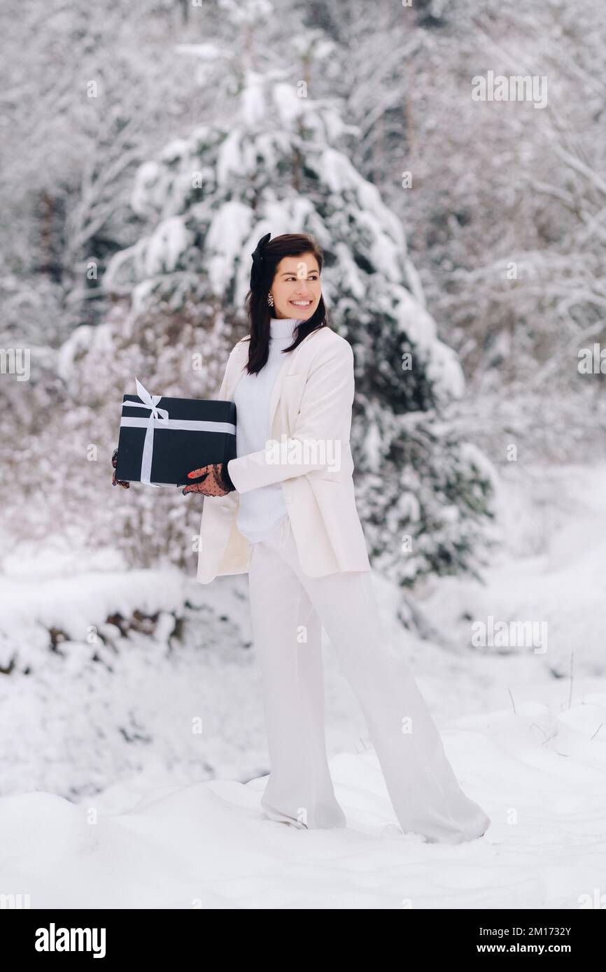 Eine stilvolle Frau mit einem weißen Anzug und einem Neujahrsgeschenk in den Händen in einem Winterwald. Ein Mädchen in der Natur in einem verschneiten Wald mit einer Geschenkbox Stockfoto