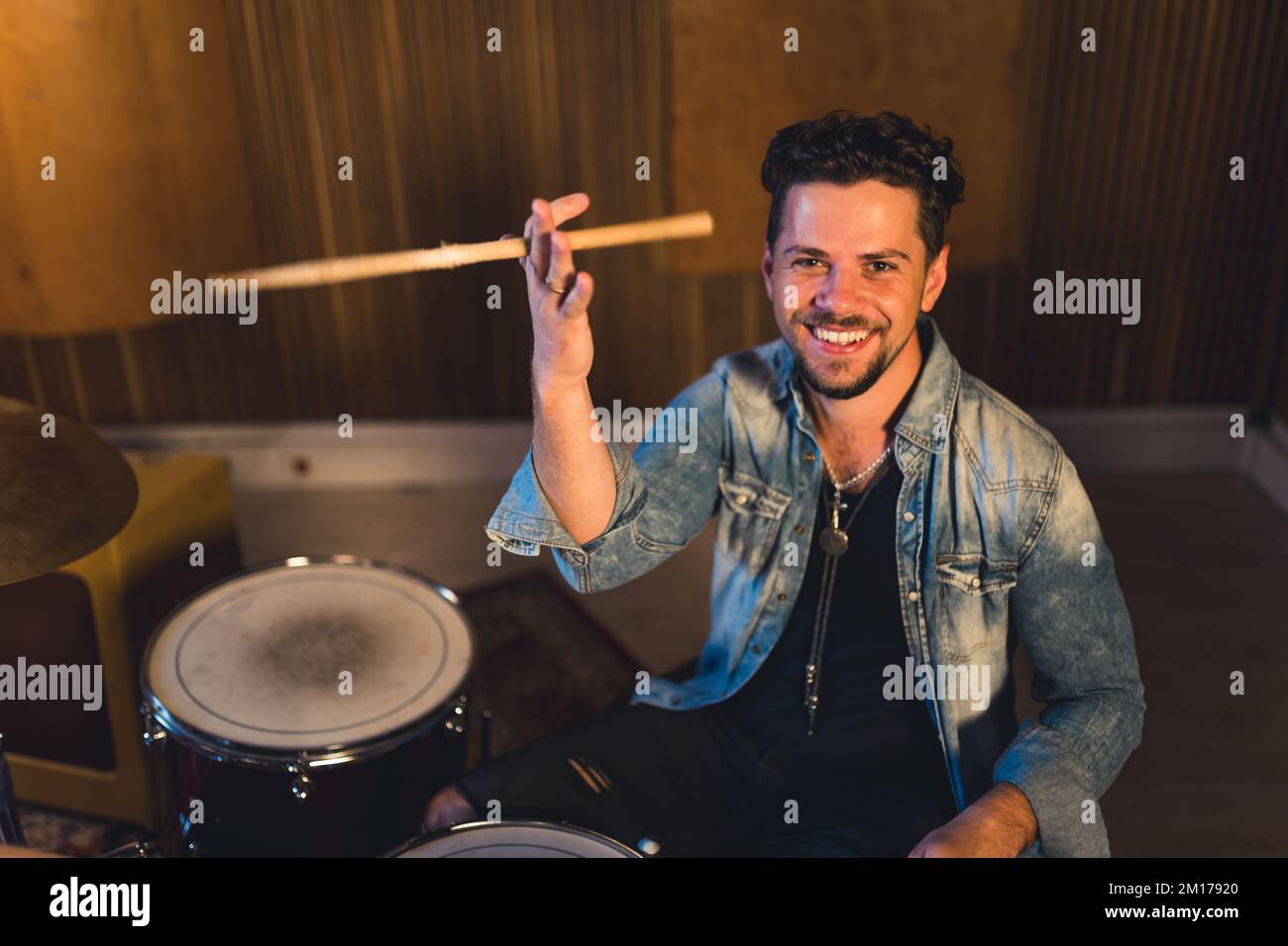 Mittellange Aufnahme eines jungen Schlagzeugers mit Schlagstöcken und einem breiten Lächeln, musikalisches Band-Konzeptstudio. Hochwertiges Foto Stockfoto