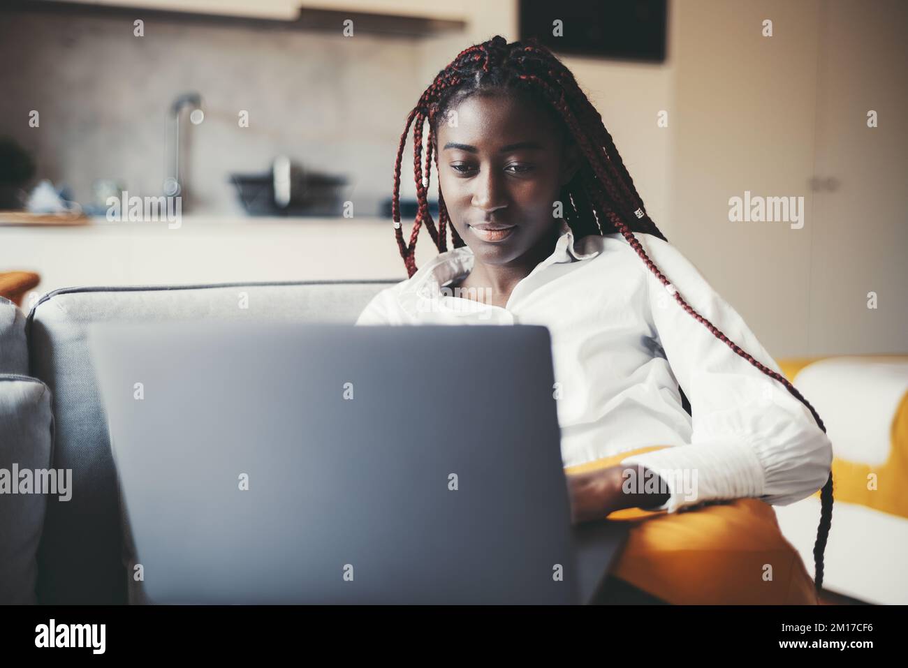 Das Porträt einer blendenden, jungen, schwarzen Freiberuflerin mit pinkfarbenen Geflechten, die an ihrem Laptop arbeiten, während sie in ihrem Wohnzimmer auf der Kutsche sitzt Stockfoto