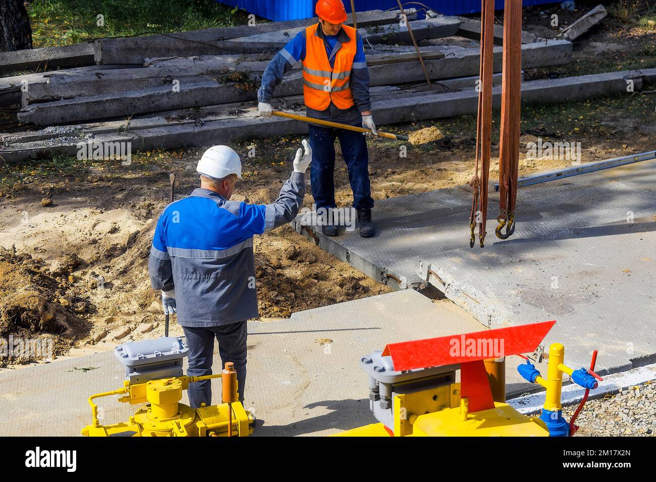 Zwei Arbeiter in Bauhelmen und Overalls, die am Sommertag Betonplatten auf der Baustelle verlegen. Bauunternehmer überwachen das Fundamentlegen auf der Baustelle. Authentischer Workflow. Stockfoto