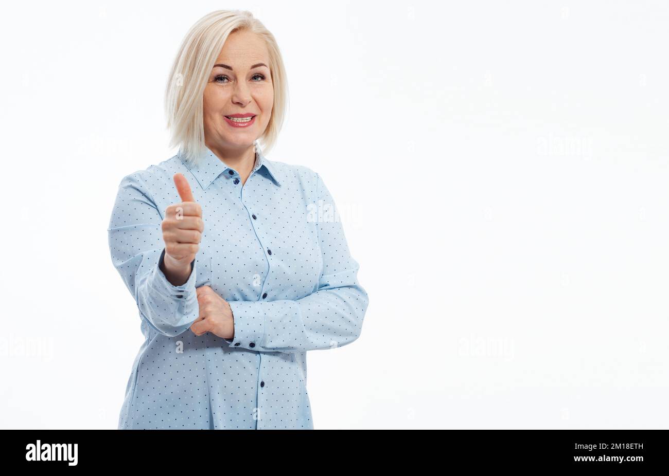 Fröhliche, lächelnde blonde Geschäftsfrau im blauen Hemd, die steht und eine gute Geste zeigt, die isoliert auf weißem Hintergrund in die Kamera schaut. Selektiver Fokus. Stockfoto