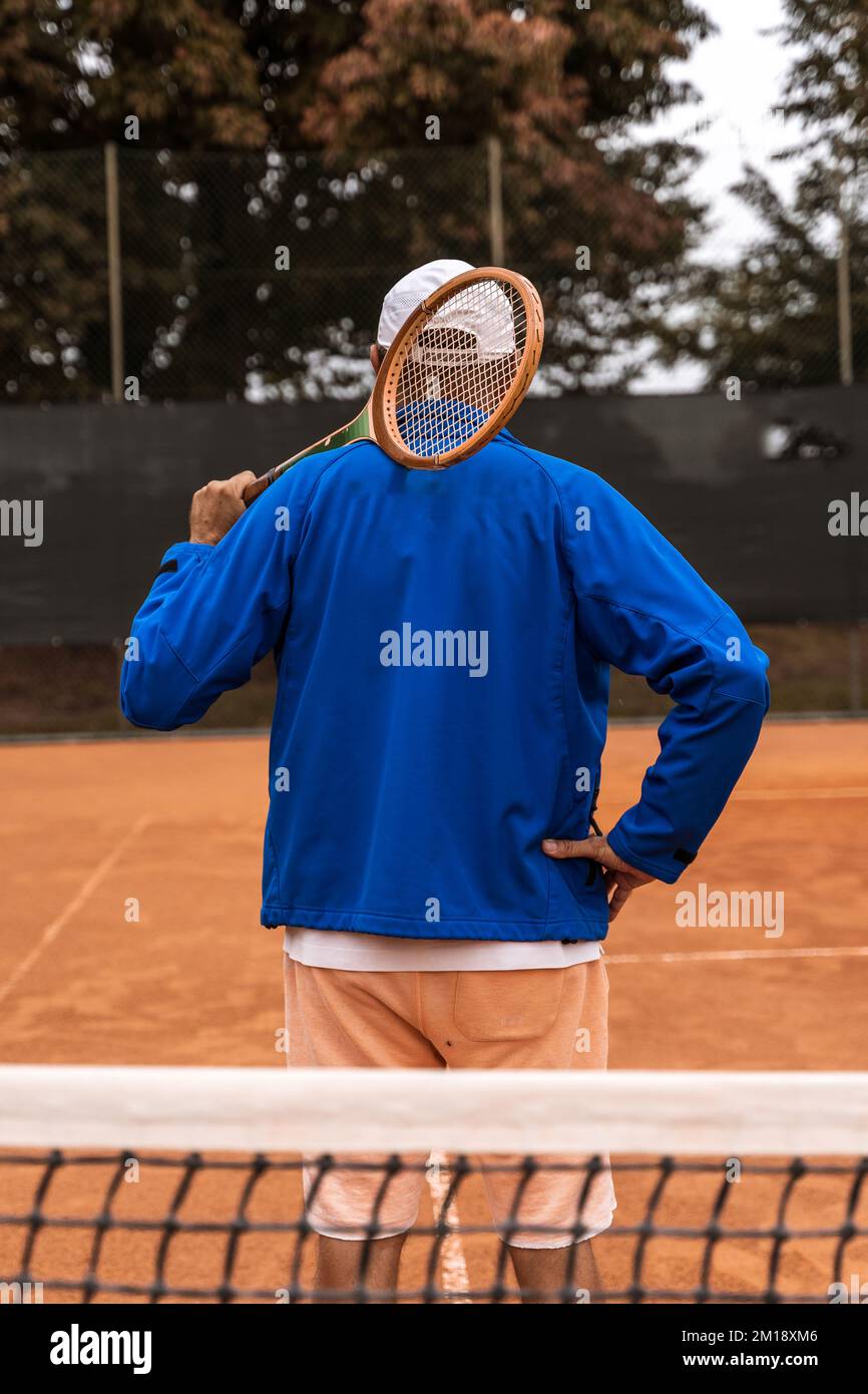 Ein pensionierter Tennisspieler in Sportbekleidung posiert mit dem Rücken zur Kamera auf einem Sandtennisplatz - Wellness-Konzept für Rentner Stockfoto