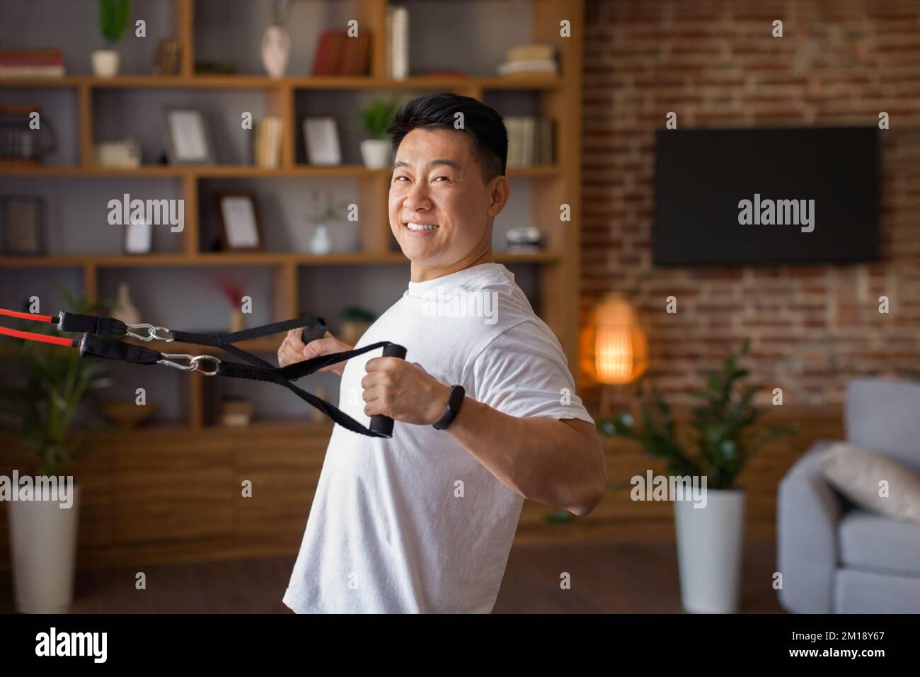 Aktiver asiatischer Mann mittleren Alters, der zu Hause mit trx-Widerstandsbändern trainiert und im Wohnzimmer trainiert Stockfoto