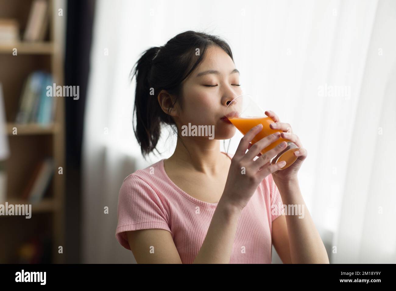 Durstige junge chinesin, die frischen Orangensaft aus Glas trinkt, steht zu Hause am Fenster, Kopierraum Stockfoto