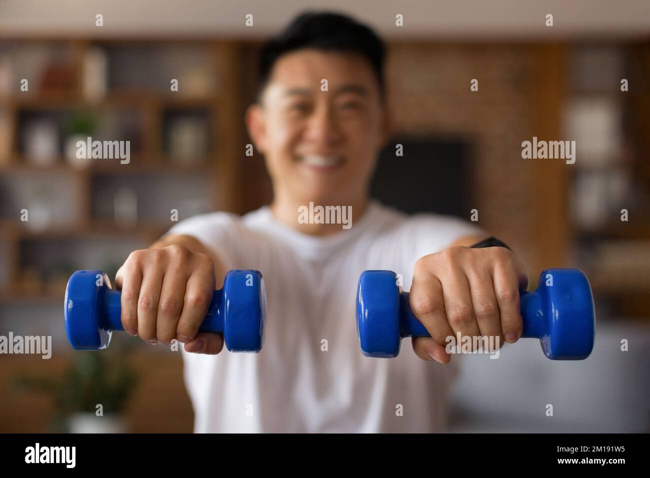 Sportlicher koreanischer reifer Mann in Sportbekleidung, der allein zu Hause trainiert, blaue Hanteln benutzt, an der Armkraft arbeitet Stockfoto