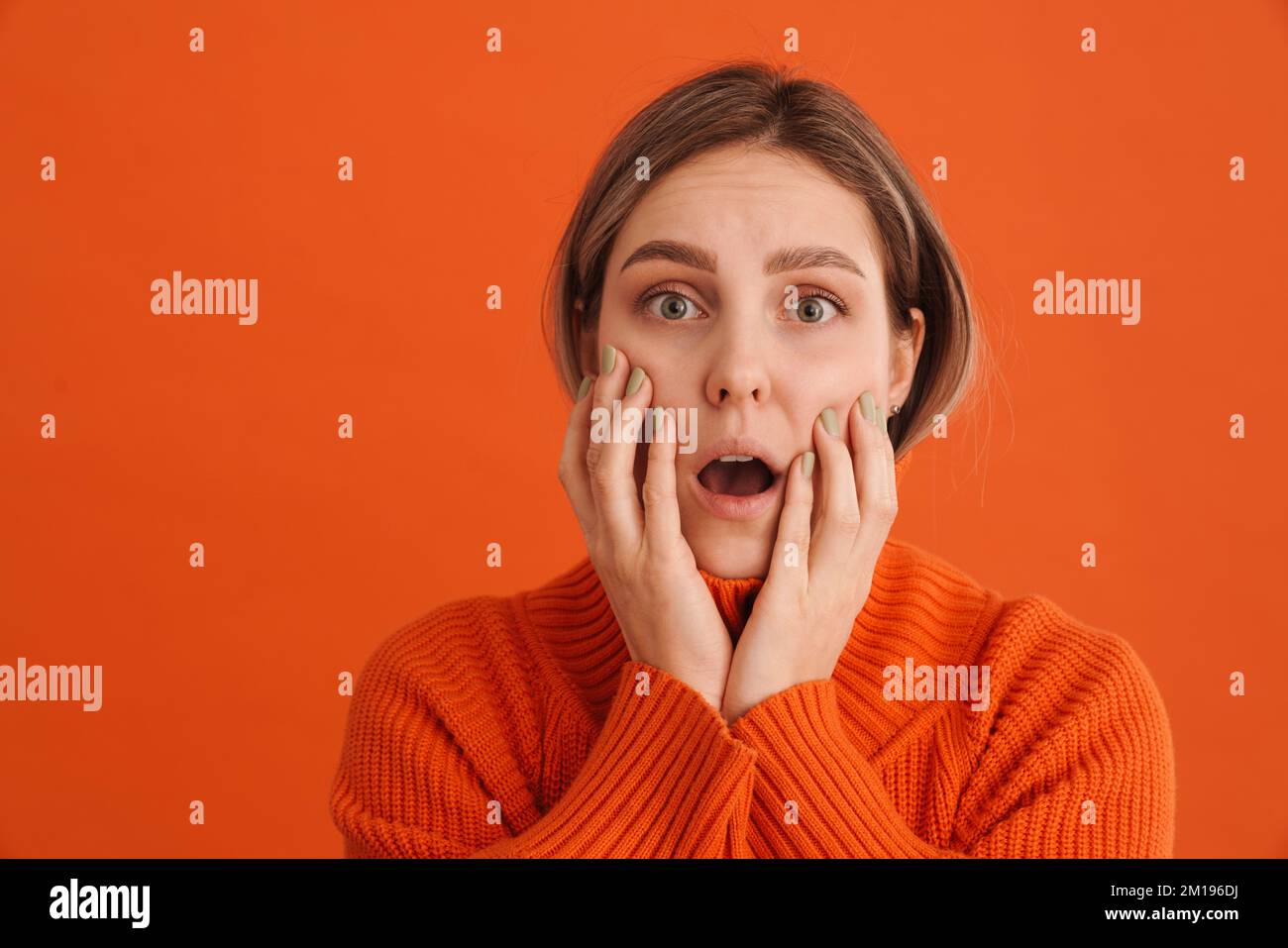 Ein junges überraschendes Mädchen, das ihre Wangen mit offenem Mund über isoliertem orangefarbenem Hintergrund berührte Stockfoto