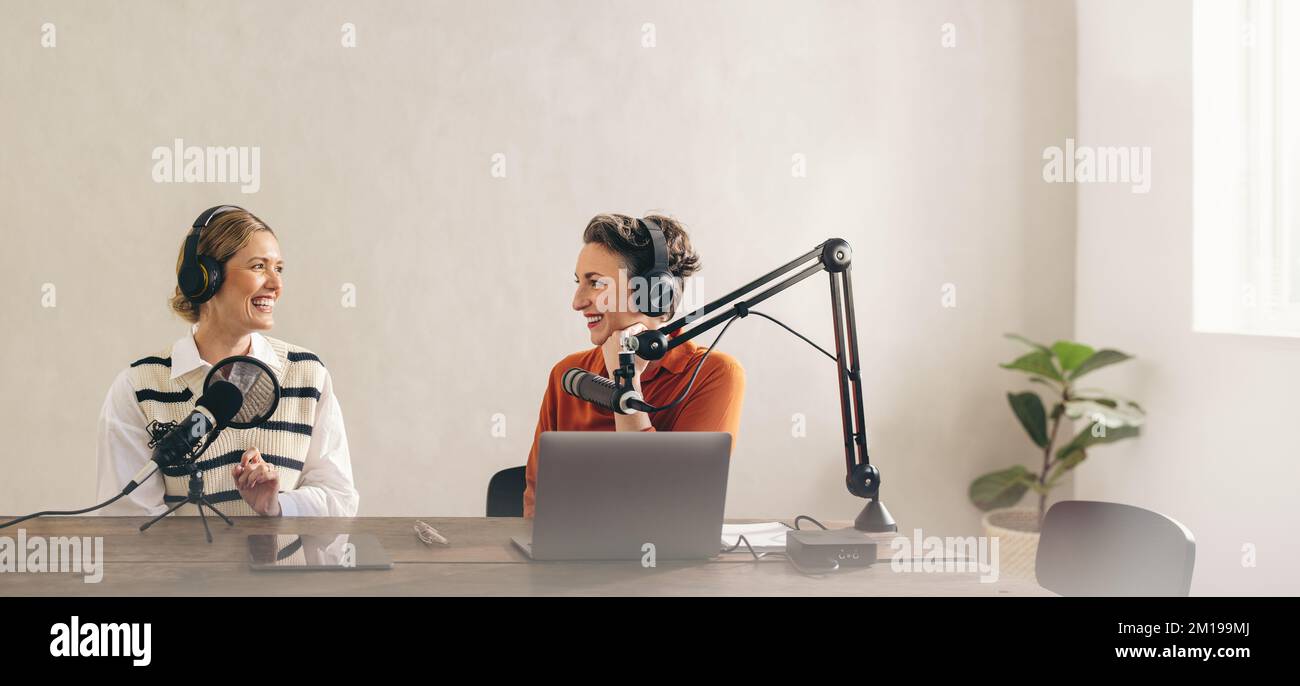 Zwei fröhliche Radio-Gastgeber, die eine großartige Unterhaltung in ihrer Sendung in einem Heimstudio führen. Zwei fröhliche Frauen lächeln, während sie einen Live-Podcast für ihren aufnehmen Stockfoto