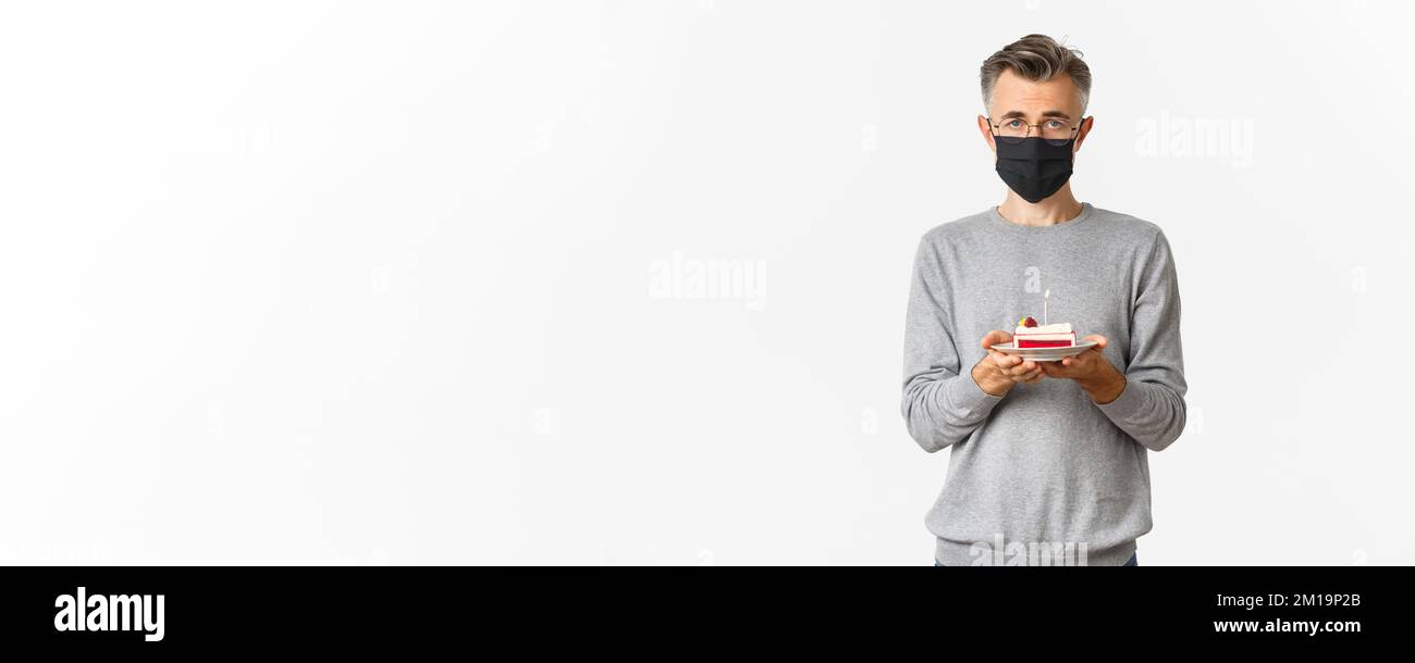Konzept von Coronavirus, Quarantäne und Urlaub. Bild von traurigen Mann mittleren Alters in schwarzer medizinischer Maske und Brille, hält Geburtstagskuchen mit brennenden Kerze Stockfoto