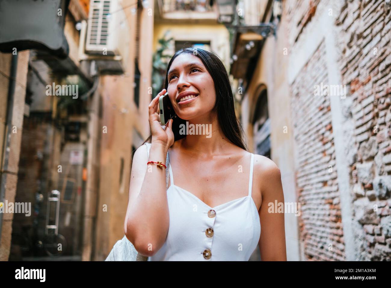 Von unten eine fröhliche lateinamerikanische Frau, die mit einem Lächeln wegsieht und die alte Straße bewundert, während sie am Wochenende in Barcelona, Spanien, ein Smartphone-Gespräch führt Stockfoto