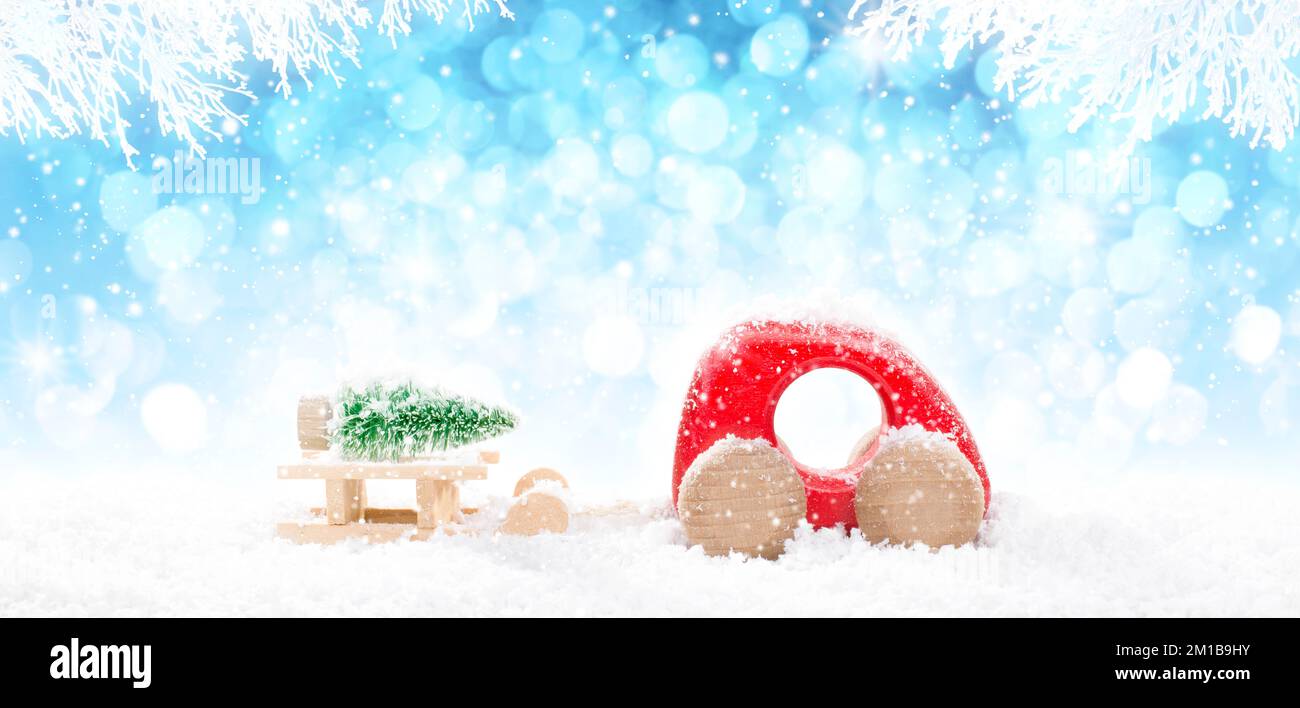 Roter hölzerner Auto-Zugschlitten mit lagengrünem Weihnachtsbaum in Perspektive auf hellblauem Bokeh-Winterhintergrund mit weißen schneebedeckten Ästen und Herbst Stockfoto