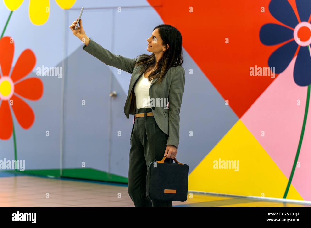 Unternehmerin in eleganter Freizeitkleidung mit lächelnder Aktentasche und Selfie an farbenfrohen Wänden mit geometrischen Verzierungen und Blumen Stockfoto