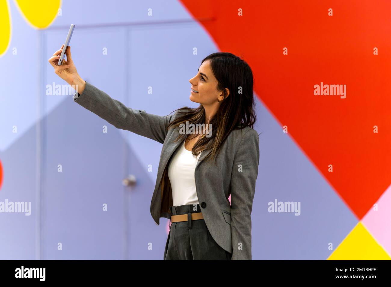 Glückliche Erwachsene Geschäftsfrau in eleganter Freizeitkleidung mit dunklen, lächelnden Haaren und Selfie an farbenfrohen Wänden am Arbeitsplatz Stockfoto