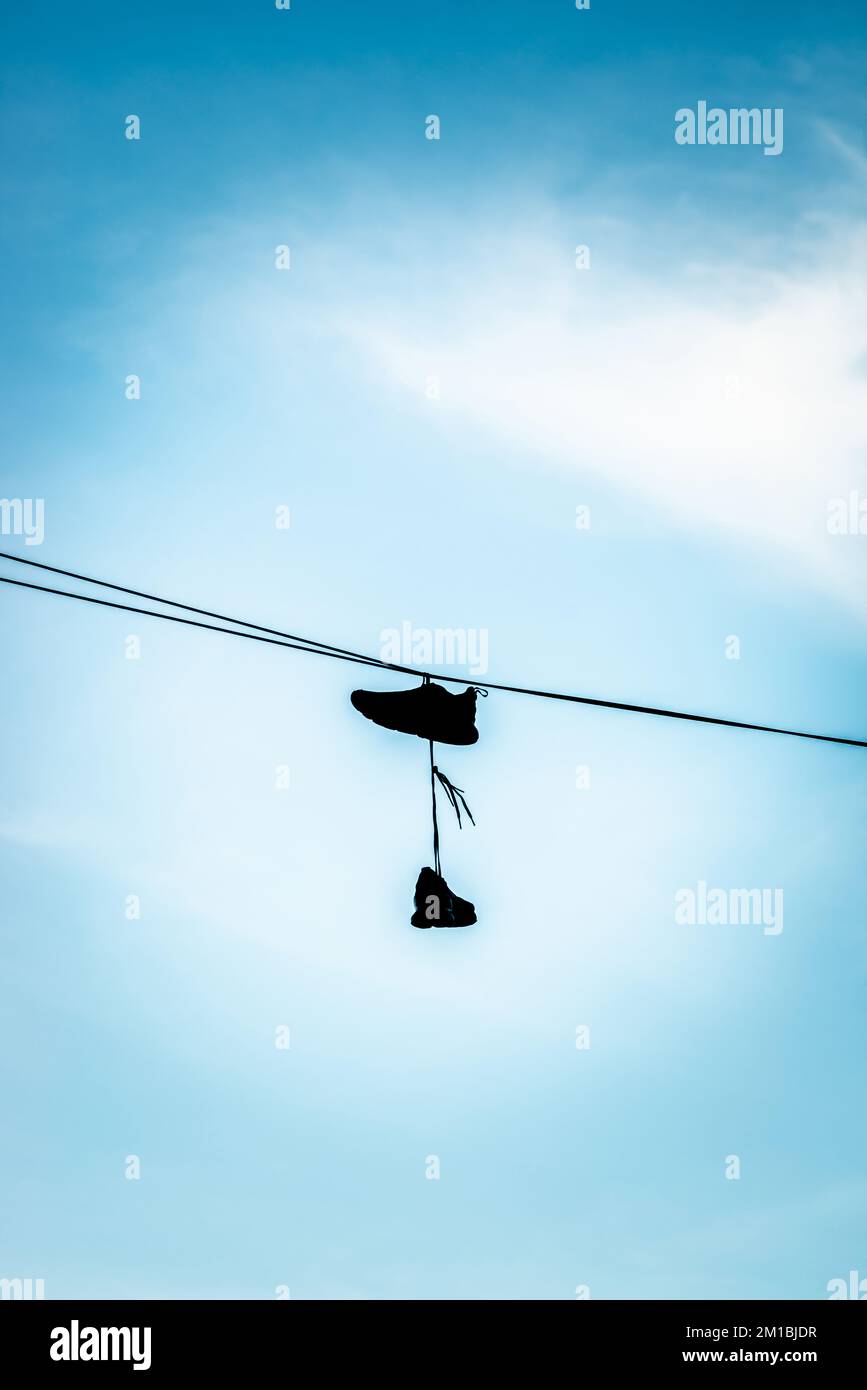 Silhouette aus aneinander gebundenen Sportschuhen, die an einer Telefonleitung vor blauem Himmel hängen, England, Großbritannien Stockfoto