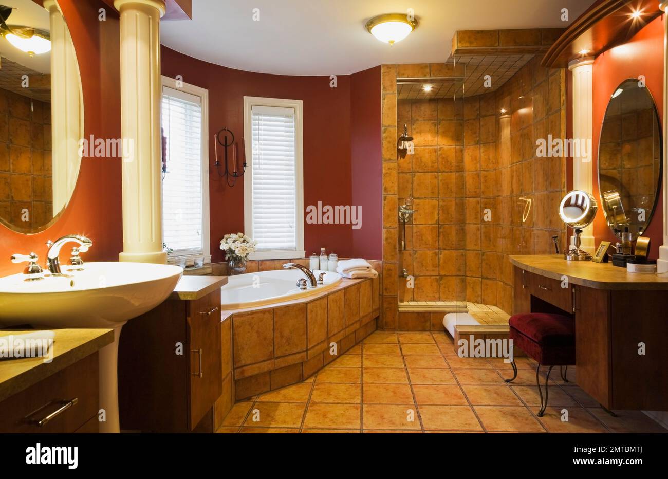 Weißes Sockelwaschbecken und Schminktisch mit roter Samtbank und Terrakotta-Keramikbadewanne im geräumigen Hauptbadezimmer im luxuriösen Haus. Stockfoto