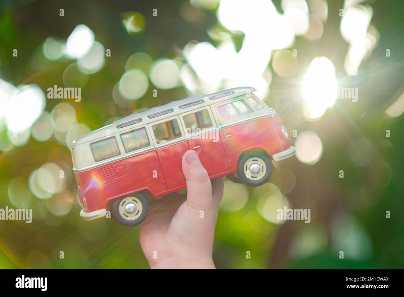 Die Hand eines kleinen Kindes hält einen roten Spielzeugwagen in Richtung Sonne. Grüner Hintergrund für den Außenbereich. Horizontaler Roadtrip, Reiseziele, Sommerurlaubskonzept. Stockfoto