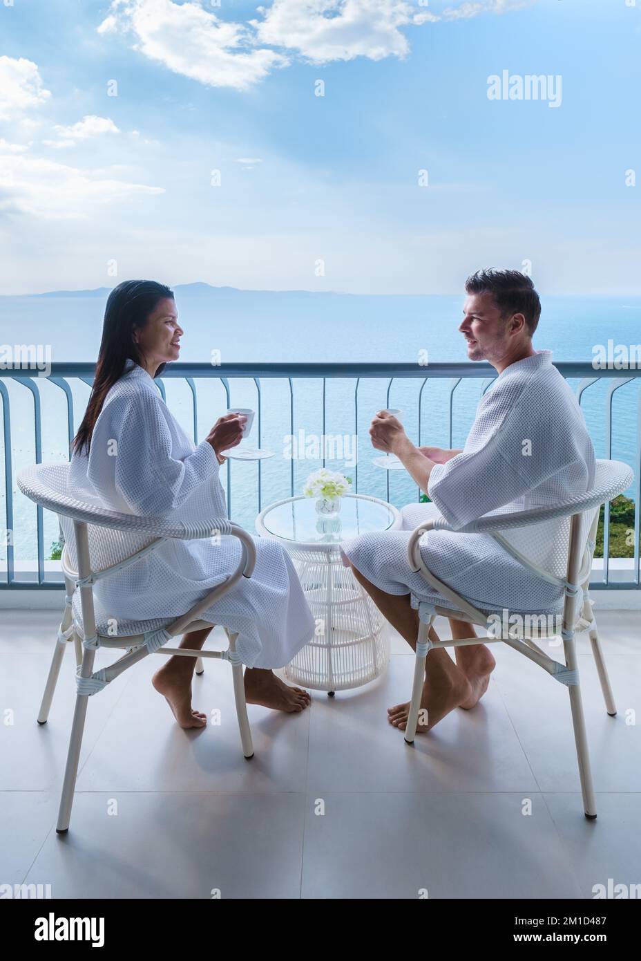 Asiatische Frauen und weiße Männer trinken Kaffee in einem Hotel auf dem Balkon mit Blick auf das Meer in Thailand Stockfoto
