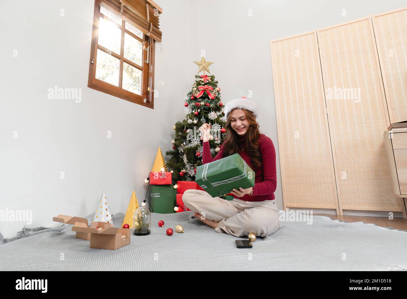 Glückliche Frau, die vor dem Hintergrund des Weihnachtsbaums sitzt. Fröhliche Dame, überrascht von der Geschenkbox. Heirate Weihnachten und frohe Feiertage Stockfoto