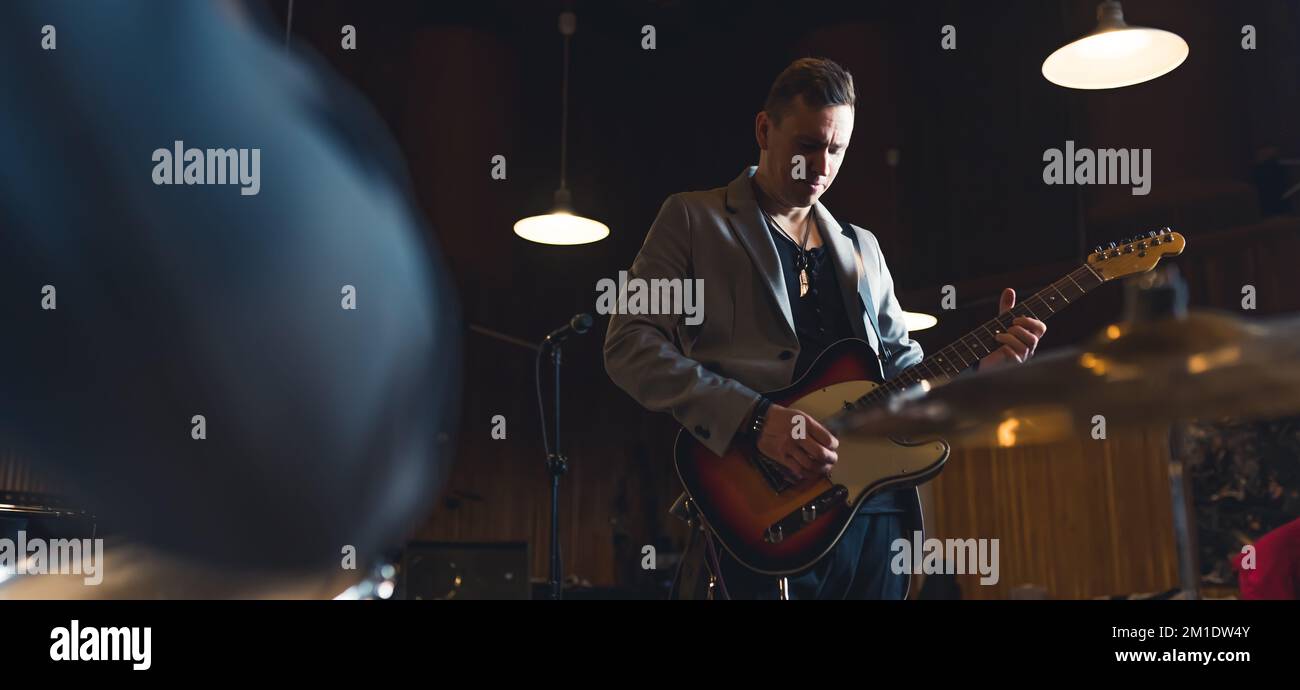 Bassist mit seiner Gitarre. Breites Innenbild eines hellhäutigen männlichen Musikers im grauen Blazer, der auf seiner Gitarre spielt. Hochwertiges Foto Stockfoto
