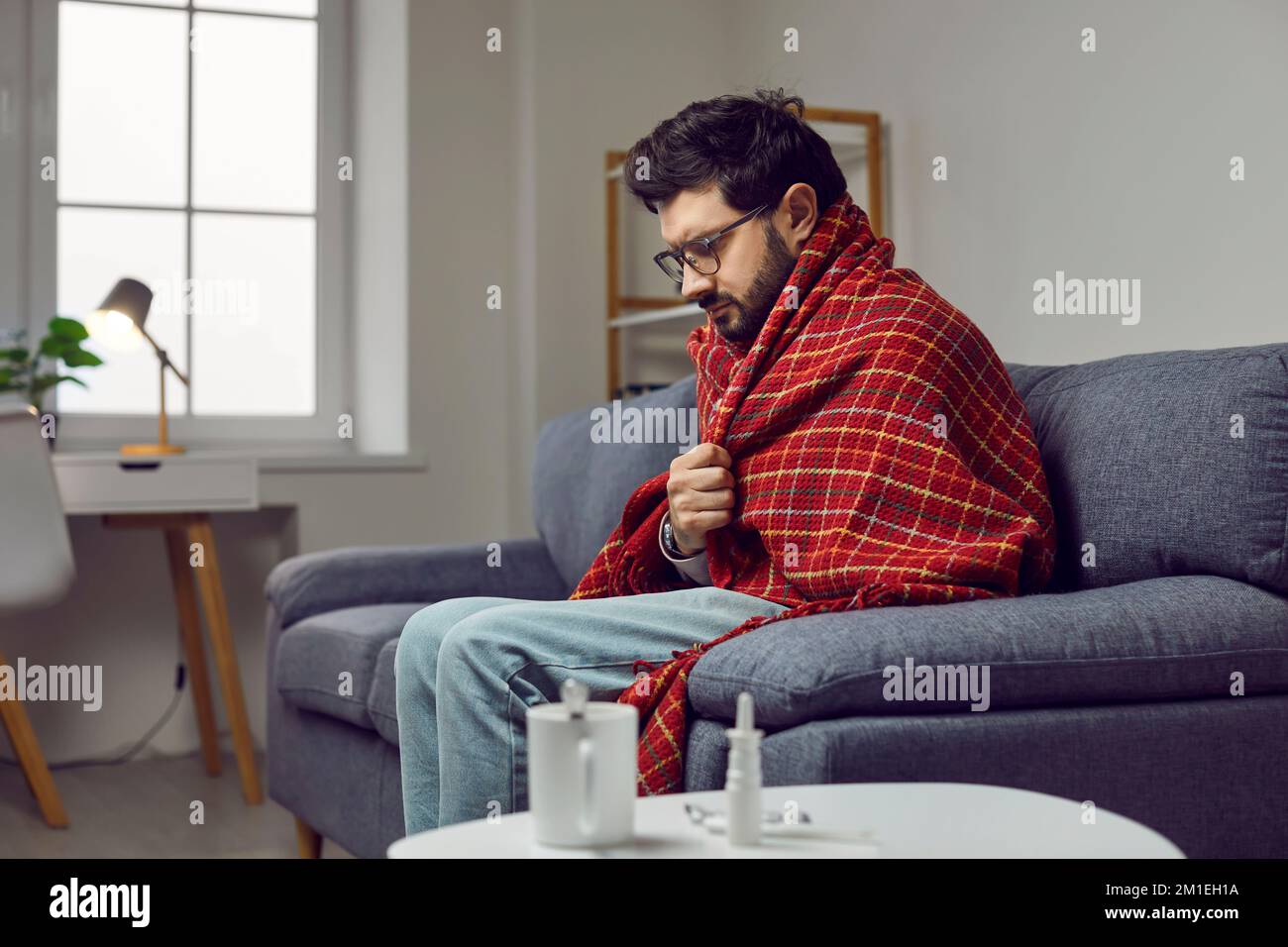 Kranker junger Mann in einer Decke, der zu Hause auf dem Sofa sitzt, sich kalt fühlt und zittert Stockfoto