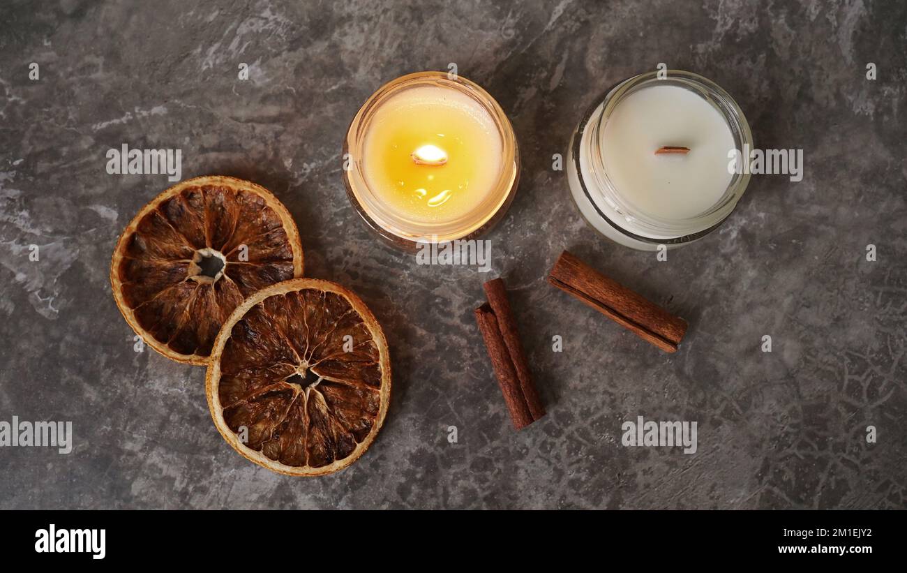 Handgefertigte brennende Kerze mit Holzdocht in weißem Glasbehälter mit Zimt und trockenen Orangen im Hintergrund, winterliche gemütliche Inneneinrichtung Stockfoto