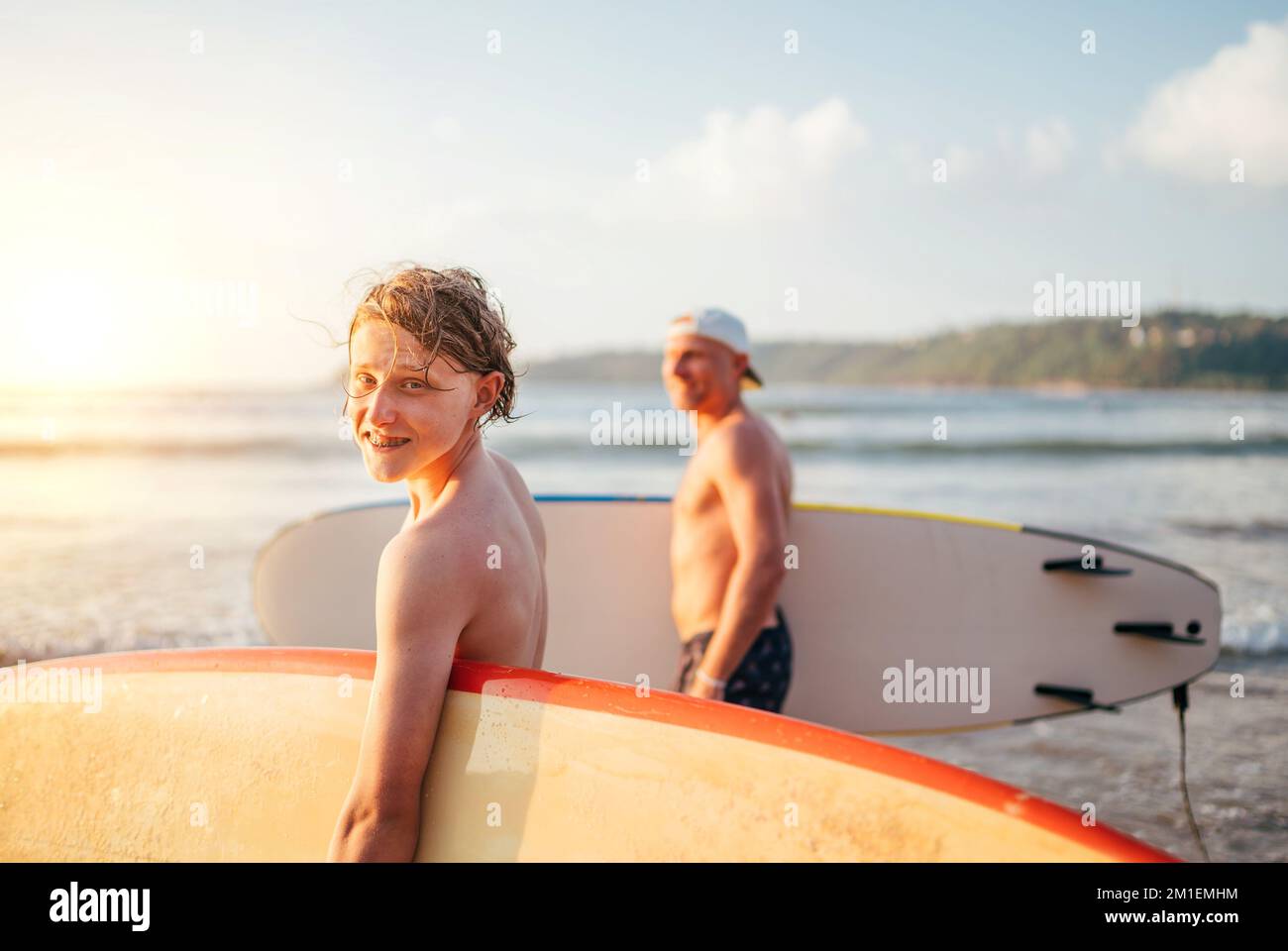 Lächelnder Teenager-Junge mit Surfbrett geht zum Surfen ins Meer. Er hat einen Winterurlaub und genießt ein schönes Sonnenuntergangslicht mit Vater auf Sri Lanka ist Stockfoto