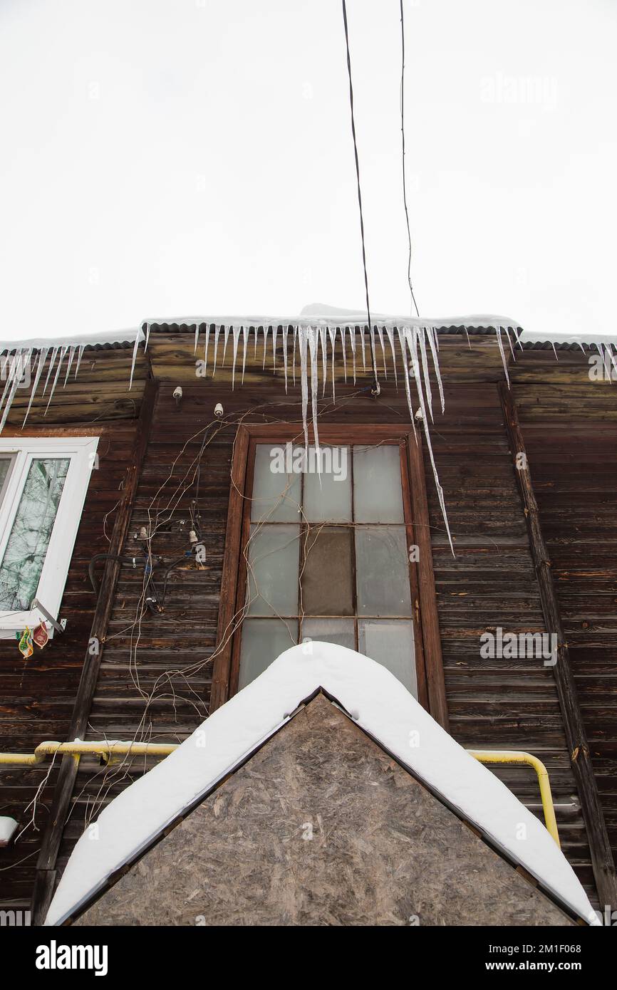 Eis, mehrere Eiszapfen hängen am Dachrand, im Winter oder Frühling. Plankenwand eines alten Holzhauses mit Fenstern. Große Eiszapfen in glatten, schönen Reihen. Bewölkter Wintertag, weiches Licht Stockfoto