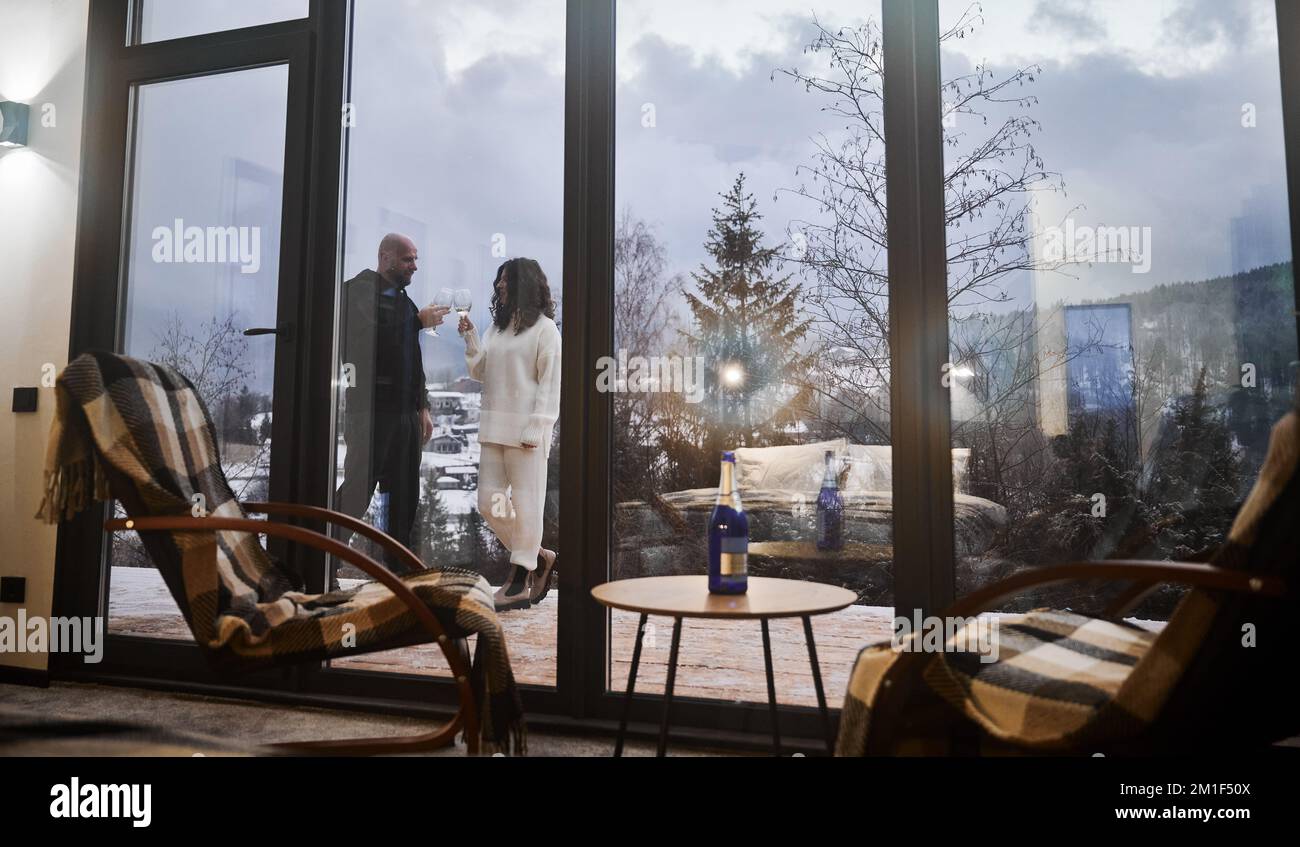 Wunderschöne Aussicht auf Mann und Frau, die mit Wein anstoßen, hinter einem Panoramafenster des skandinavischen Barnhouse. Die Inneneinrichtung des Holzhauses mit Stühlen und einem Paar, das sich außerhalb des Gebäudes verliebt hat. Stockfoto