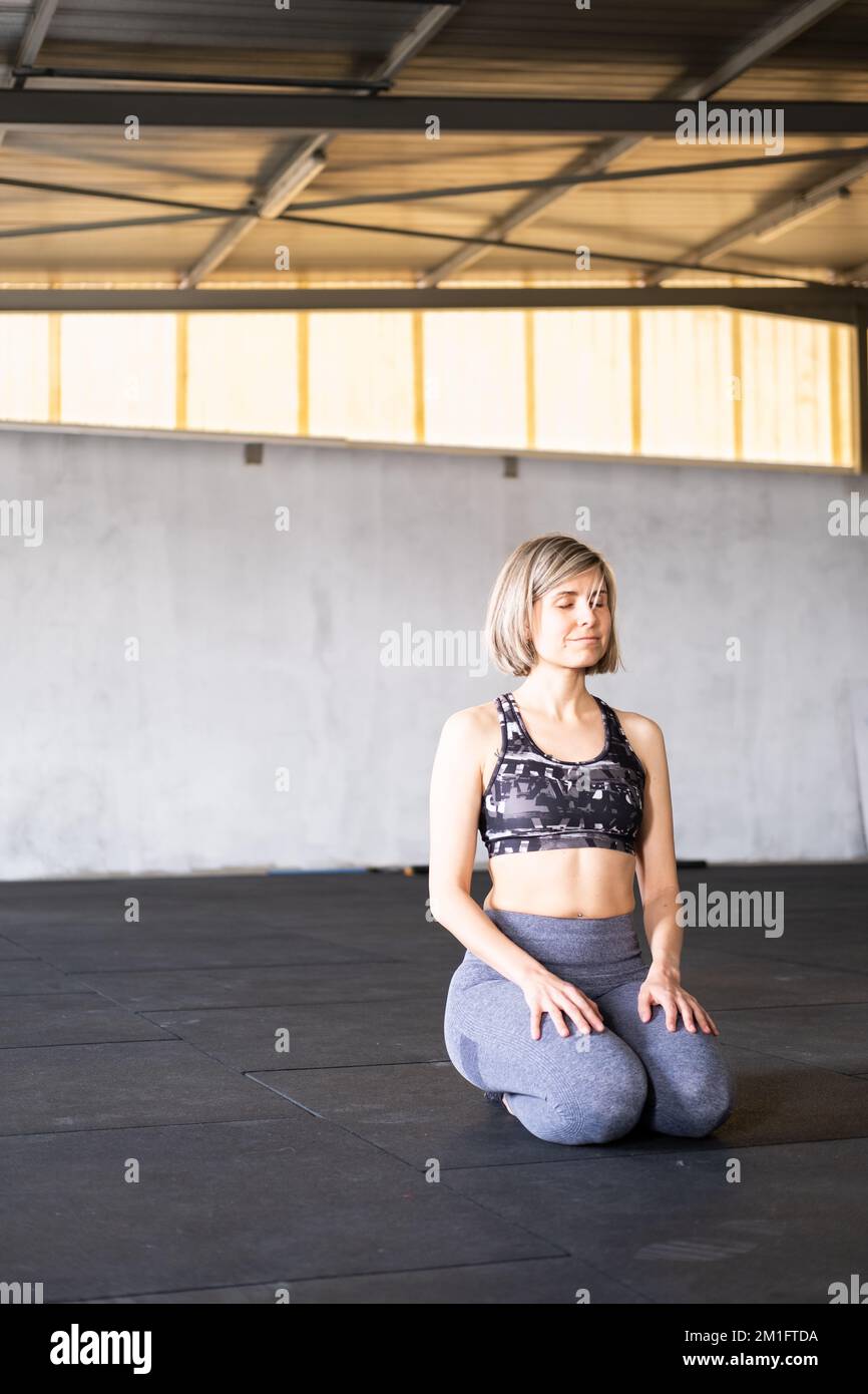 Vertikales Ganzkörperfoto einer Smiley-Mid-Adult-Frau, die sich im Knien ausruht und meditiert, oder vajrasana-Pose mit geschlossenen Augen während ihres Vinyasa-Flusses Stockfoto