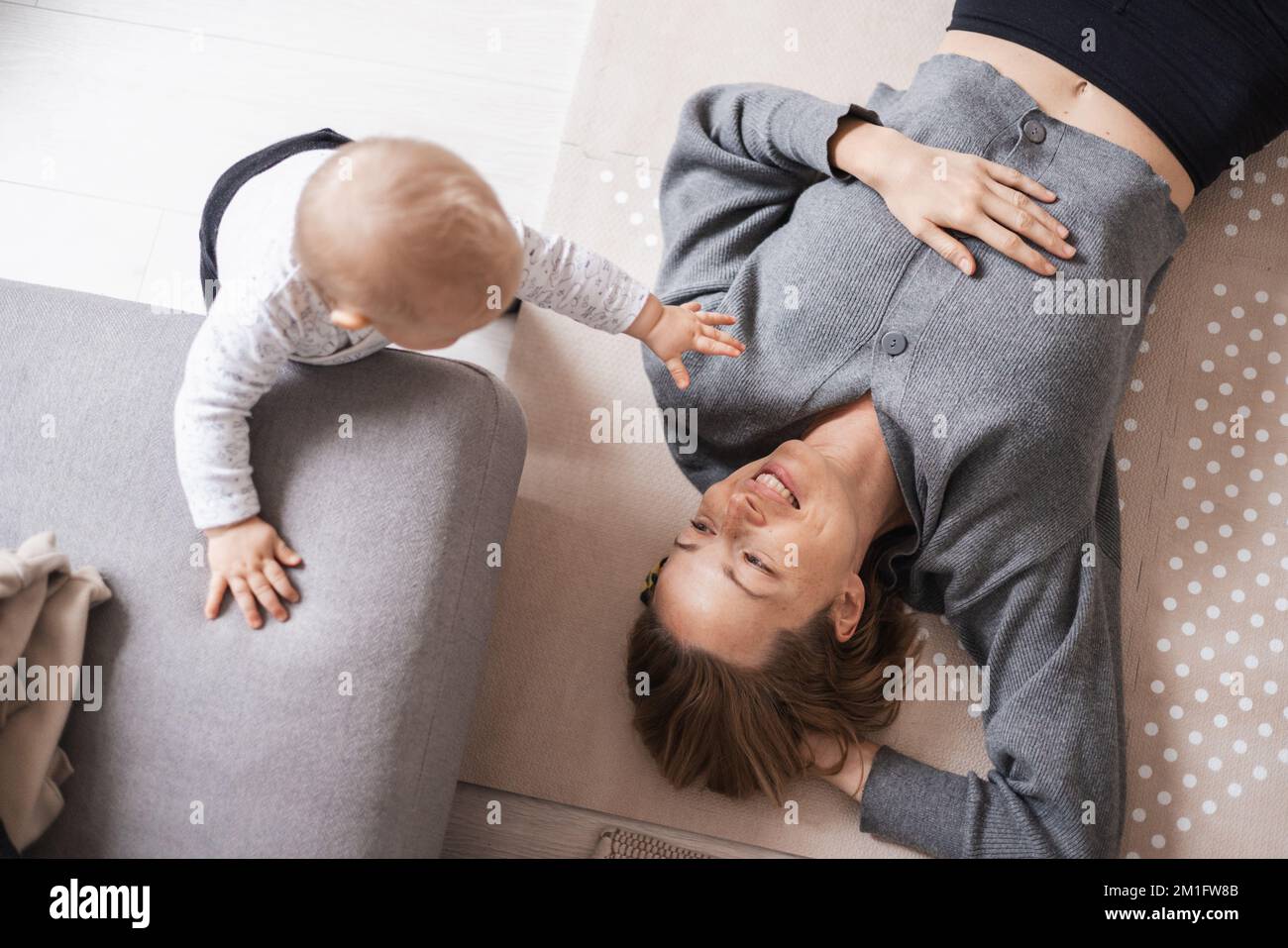 Glückliche Familienmomente. Die Mutter liegt bequem auf der Kindermatte und spielt dabei mit ihrem kleinen Jungen, der seine ersten Schritte beobachtet und überschwemmt. Positive menschliche Emotionen, Gefühle, Freude Stockfoto