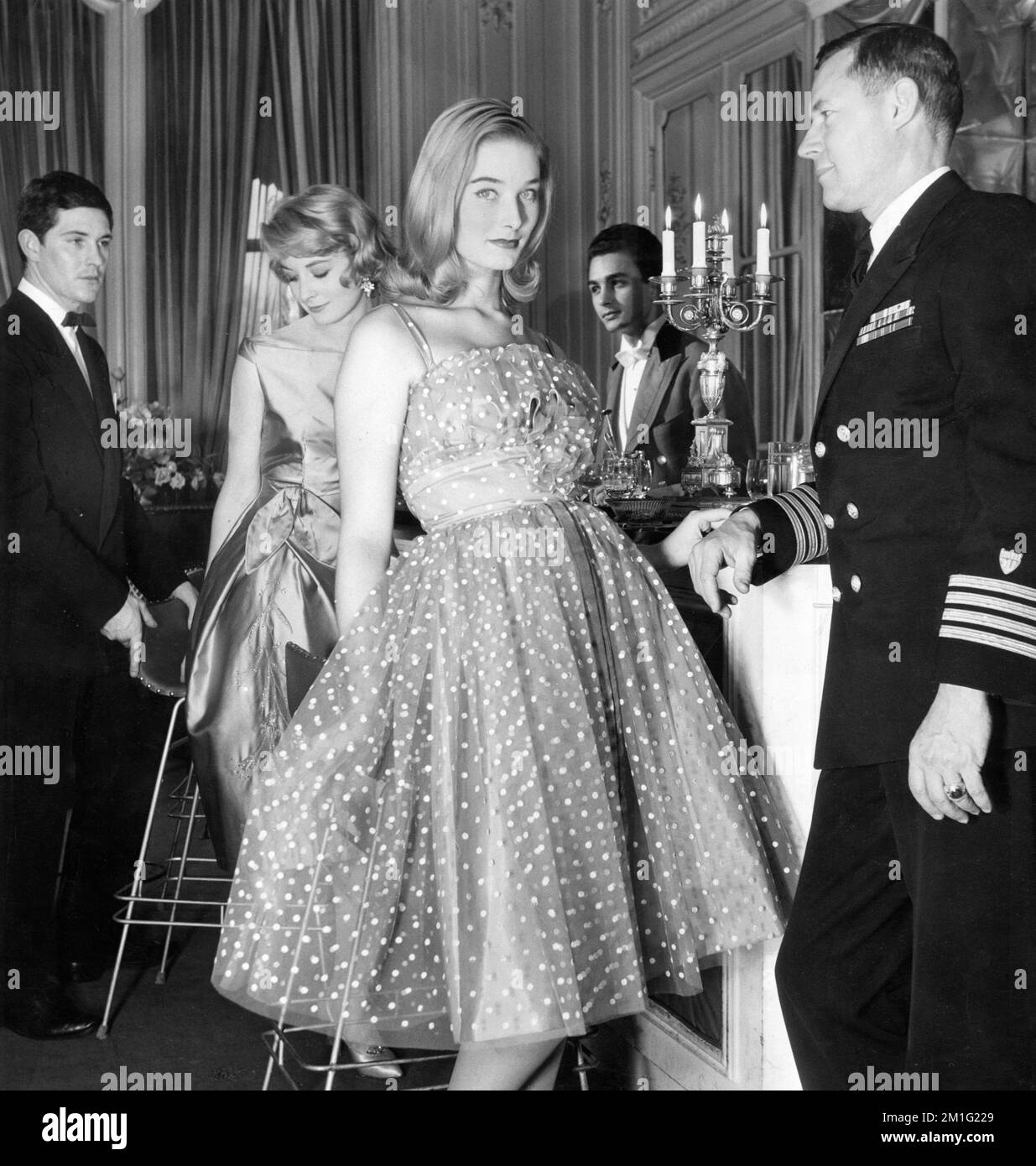 Modeartikel der englischen Modemodelle SANDRA PAUL in Cocktailkleid und TANIA MALLETT in einem kurzen Abendkleid mit dem Kleiderdesigner DAVID SKINNER links und Captain RUSSELL R. WAESCHE von der US-Küstenwache, stationiert in London rechts, fotografiert im Helene Cordet's Dining Club um November 1958 Stockfoto