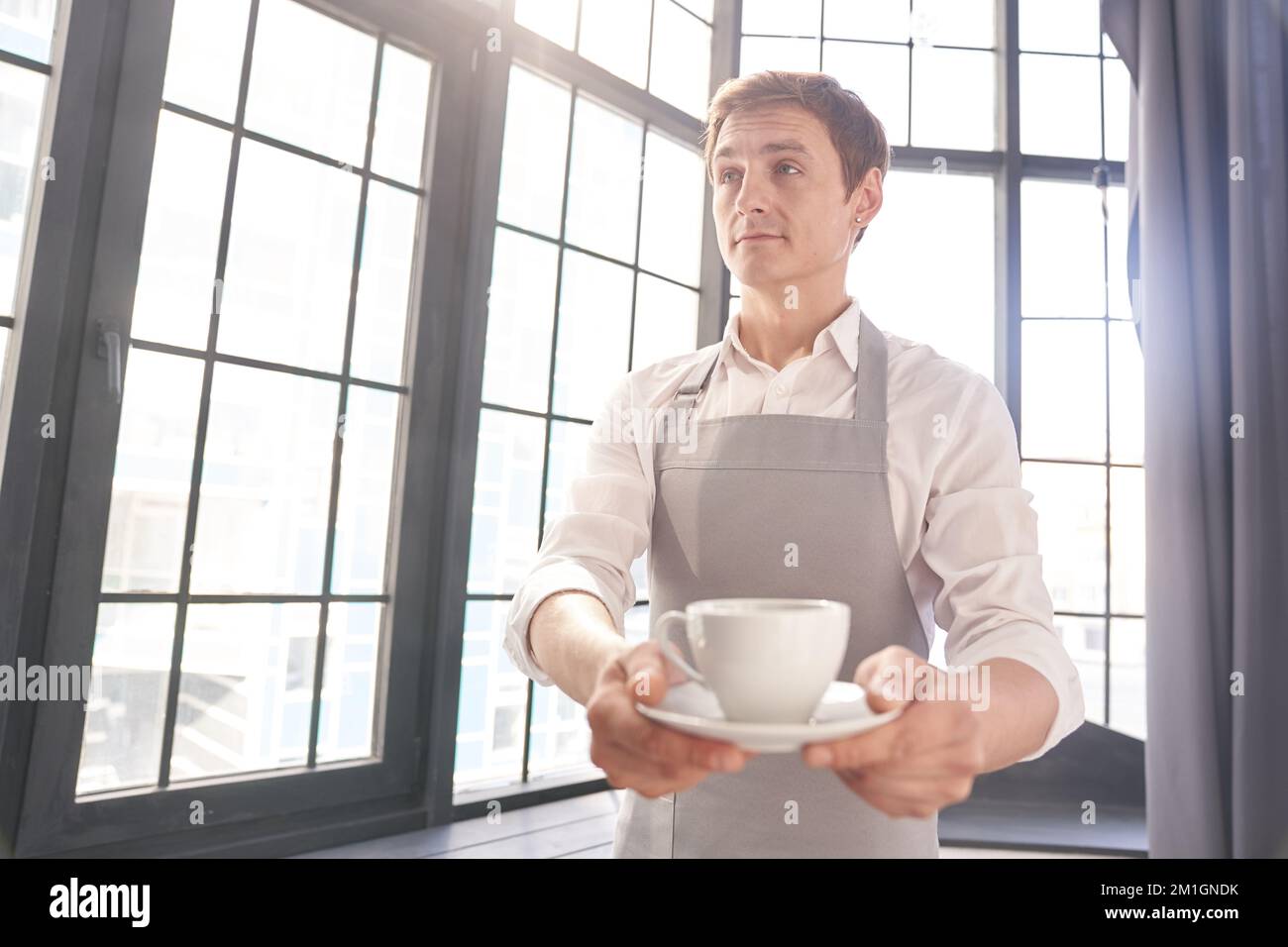 Ein männlicher Kellner blickt in einer grauen Schürze weg und hält eine Tasse Kaffee aus. Der Barista serviert eine Tasse heißen Kaffee vor dem Hintergrund eines großen Fensters mit Platz zum Kopieren. Hochwertiges Foto Stockfoto