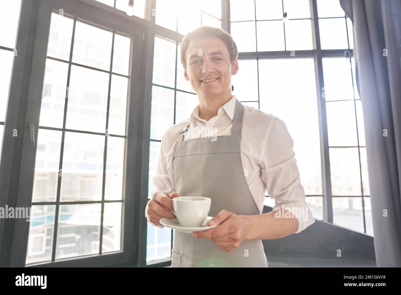 Ein Kellner in einer grauen Schürze hält eine Tasse Kaffee. Der Barista gibt eine Tasse heißen Kaffee in einem Café vor dem Hintergrund eines großen Fensters. Hochwertiges Foto Stockfoto