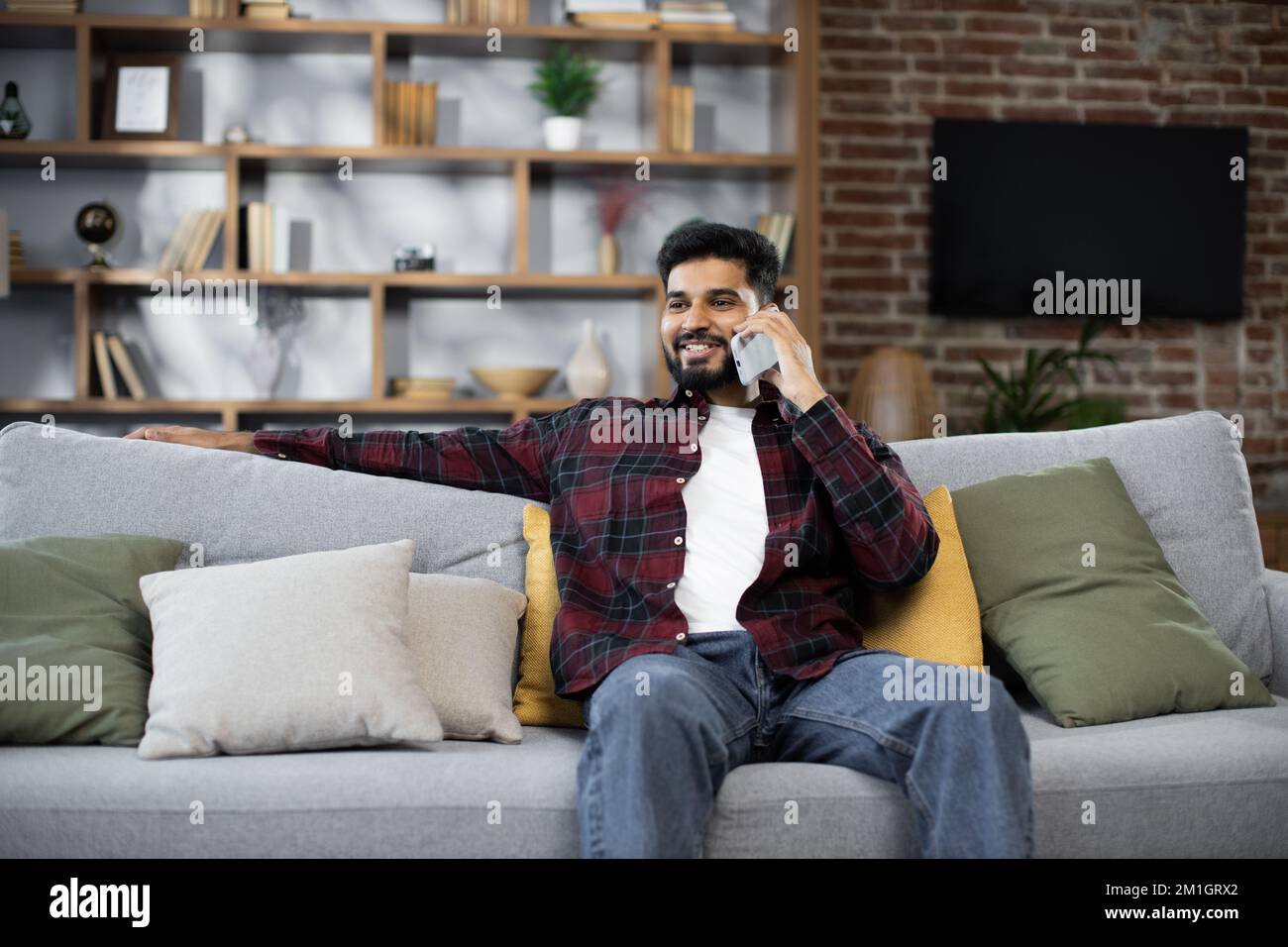 Gutaussehender, bärtiger, positiver Mann, der auf der Couch sitzt und seinen Freund mit dem Smartphone im Haus anruft. Lächelnder männlicher Freiberufler, der während der Heimarbeit Pause macht. Stockfoto