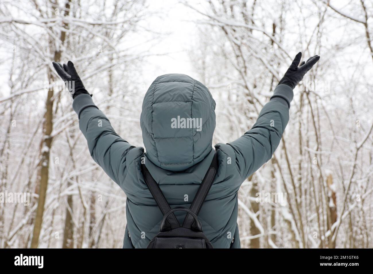 Eine Frau trägt eine Daunenjacke mit Kapuze, steht mit erhobenen Händen und genießt das schneebedeckte Wetter. Freizeit im Winterpark, kalte Jahreszeit Stockfoto