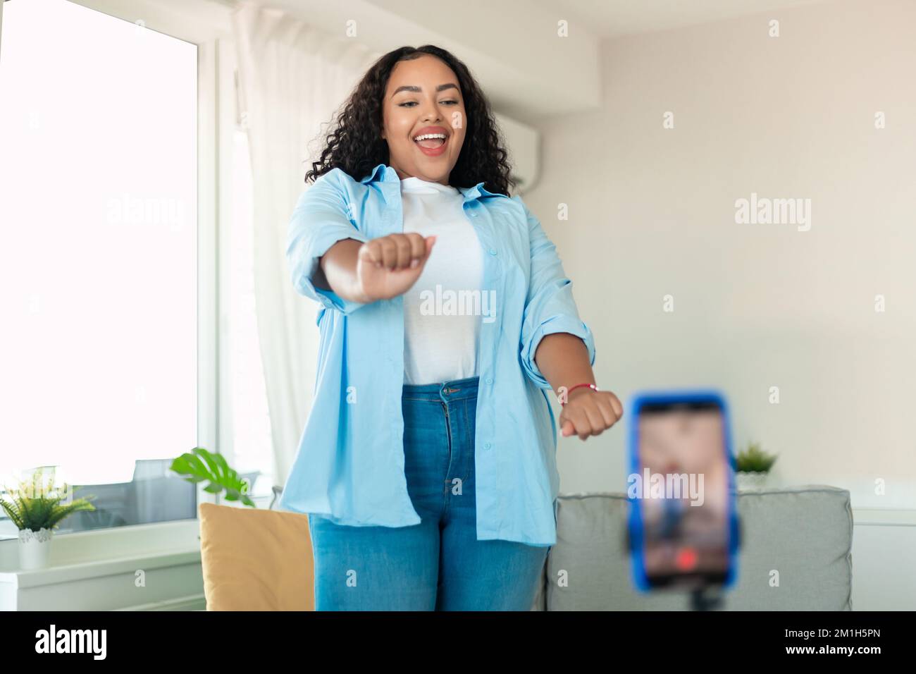 Fröhliche, schwarze, dicke Frau, die in der Handykamera tanzt und zu Hause Videos mit dem Telefon auf dem Stativ filmt Stockfoto