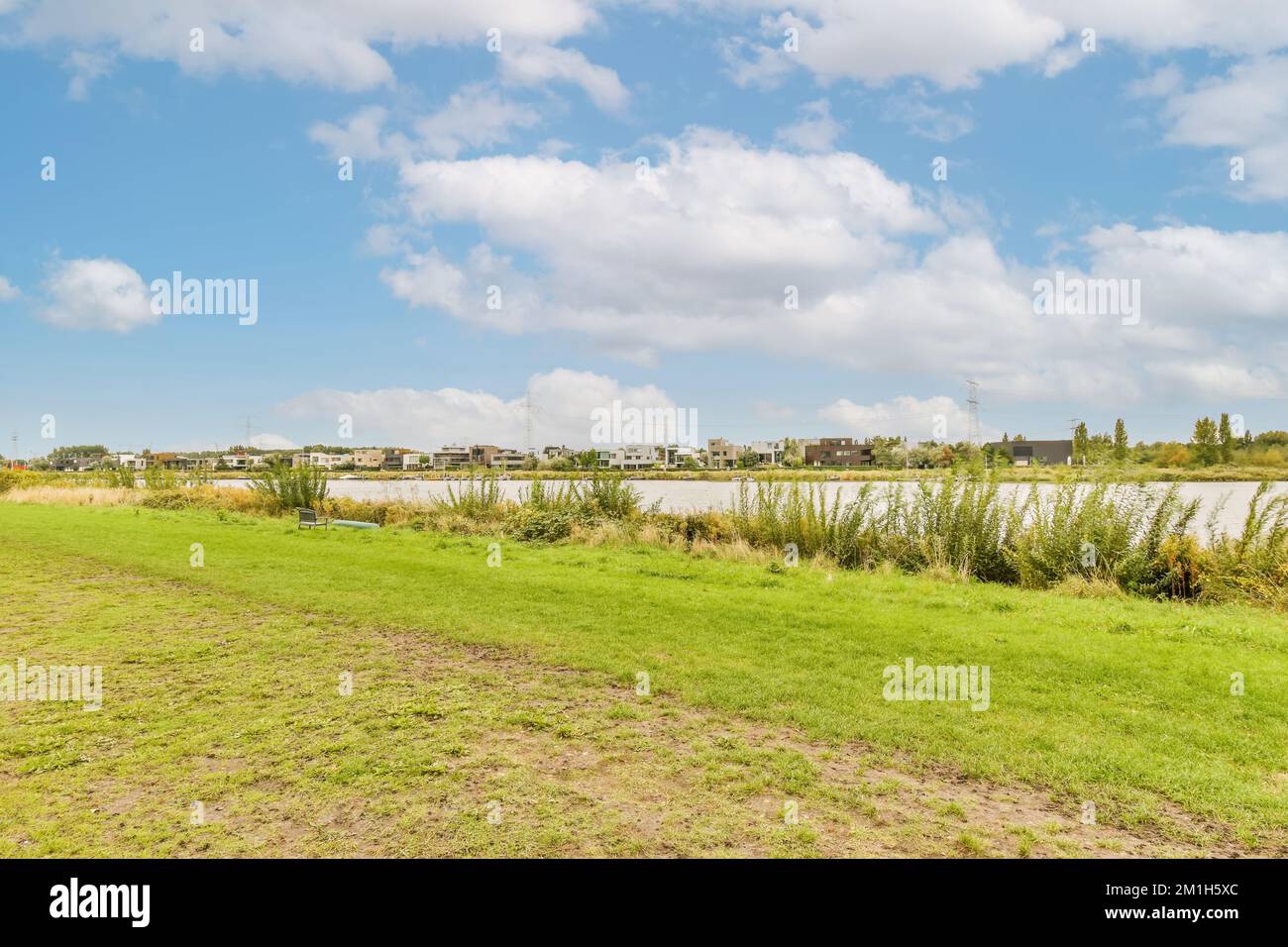 Ein grasiges Feld mit Wasser im Hintergrund und einige Gebäude auf dem anderen Land zum Himmel sind teilweise blau Stockfoto