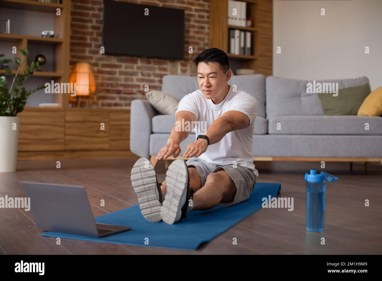 Heimtrainingkonzept. Aktiver, reifer asiatischer Mann, der sich die Beinmuskulatur vor dem Laptop streckt und sich online Videoanleitungen ansieht Stockfoto