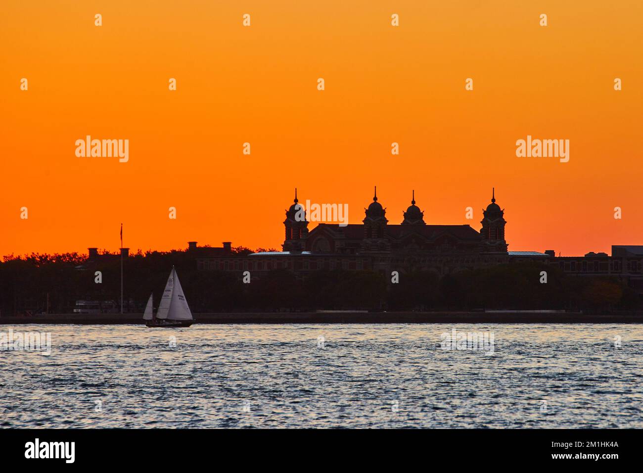 Silhouette von Ellis Island mit vorbeisegelndem Boot und intensivem orangefarbenen Himmel Stockfoto