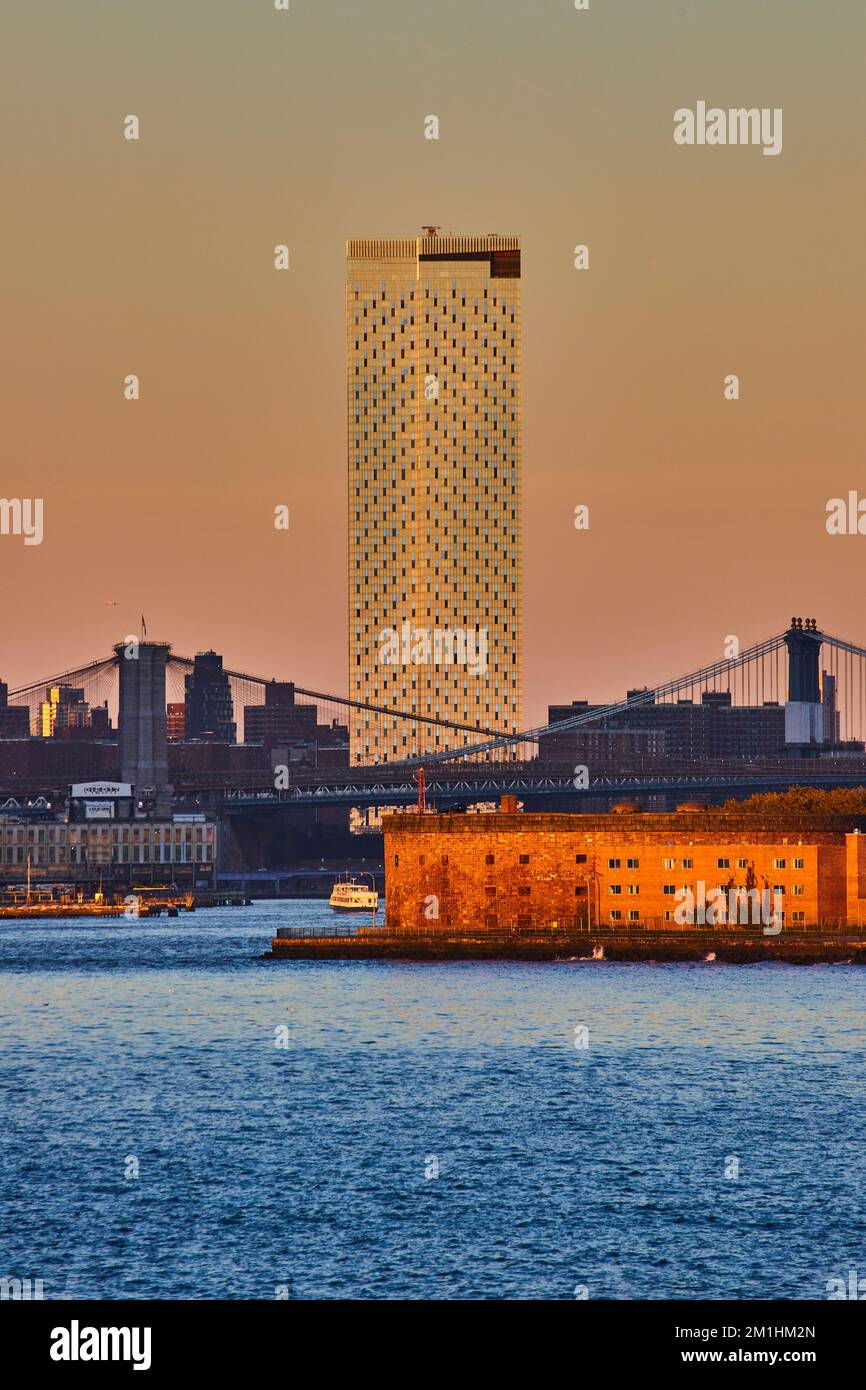 Einsamer Wolkenkratzer und Governors Island mit Manhattan und Brooklyn Bridges vom Wasser aus in der goldenen Stunde von New York City Stockfoto