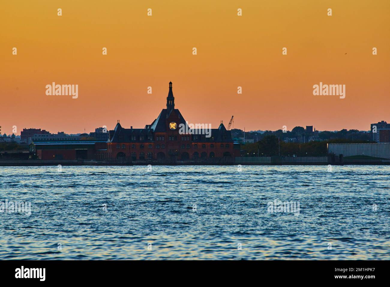 New Jersey Fährstation nach Ellis Island von Wasser mit glühender Uhr und orangefarbenem Dämmerlichthimmel Stockfoto