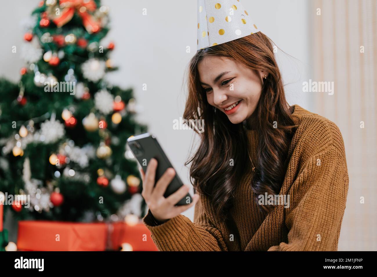 Fröhliche Frau, die die Weihnachtsbotschaft liest, Glückwunsch Nachricht Benutzen Sie Ihr Smartphone an einem festlich dekorierten Weihnachtsbaum. Lächelnde, wunderschöne Frau, die sich auf dem Handy unterhält Stockfoto