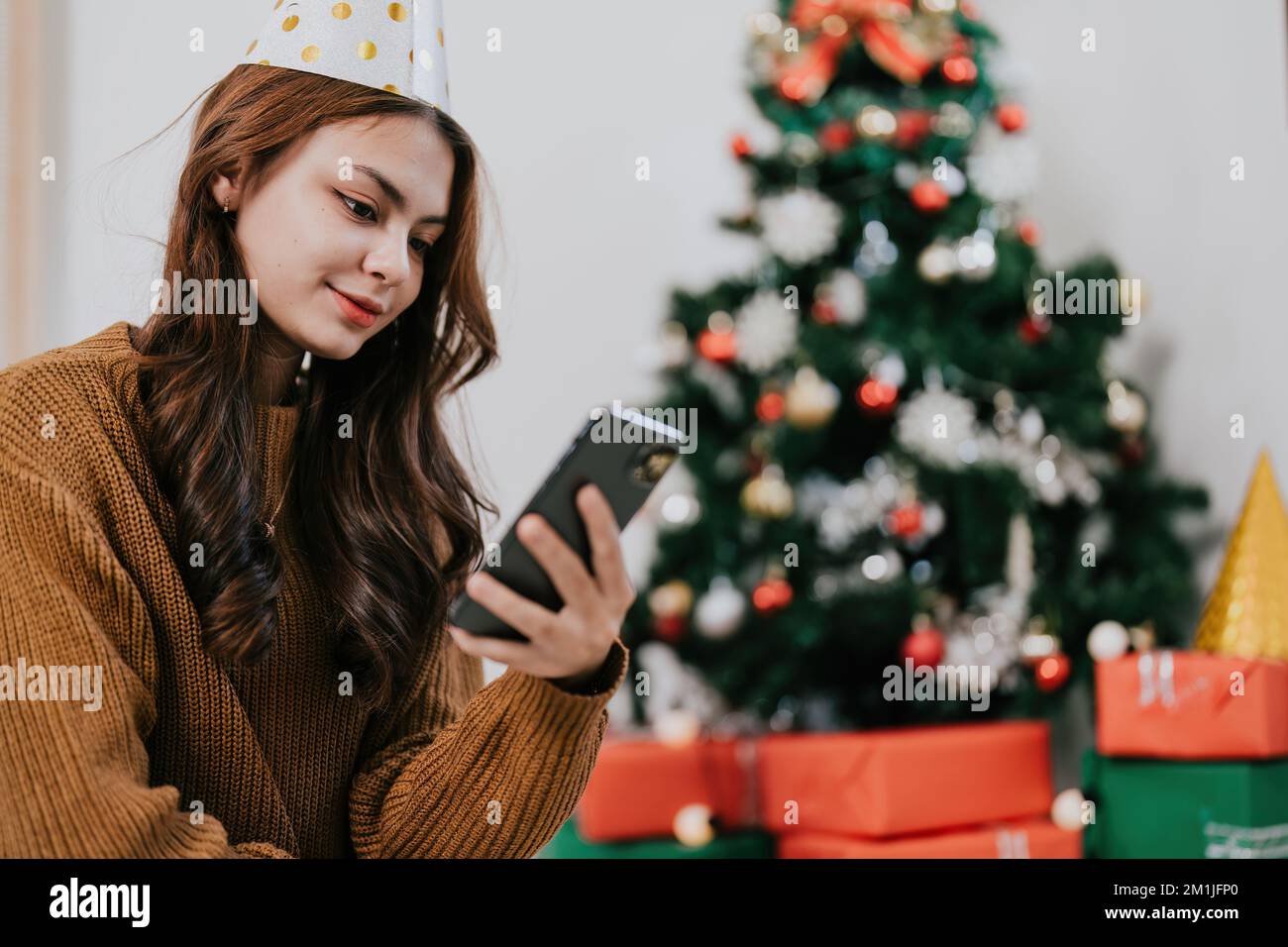 Fröhliche Frau, die die Weihnachtsbotschaft liest, Glückwunsch Nachricht Benutzen Sie Ihr Smartphone an einem festlich dekorierten Weihnachtsbaum. Lächelnde, wunderschöne Frau, die sich auf dem Handy unterhält Stockfoto