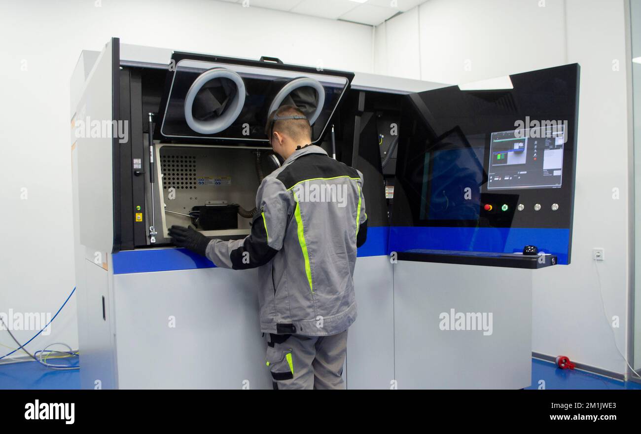 Ein Arbeiter blickt in ein offenes Fenster eines großen industriellen 3D-Druckers für Metallpulver. Der Arbeitsprozess. Industrial Powder 3D Drucker für Metall und Arbeiter Mann. Laser-Sintermaschine für Metall. Stockfoto
